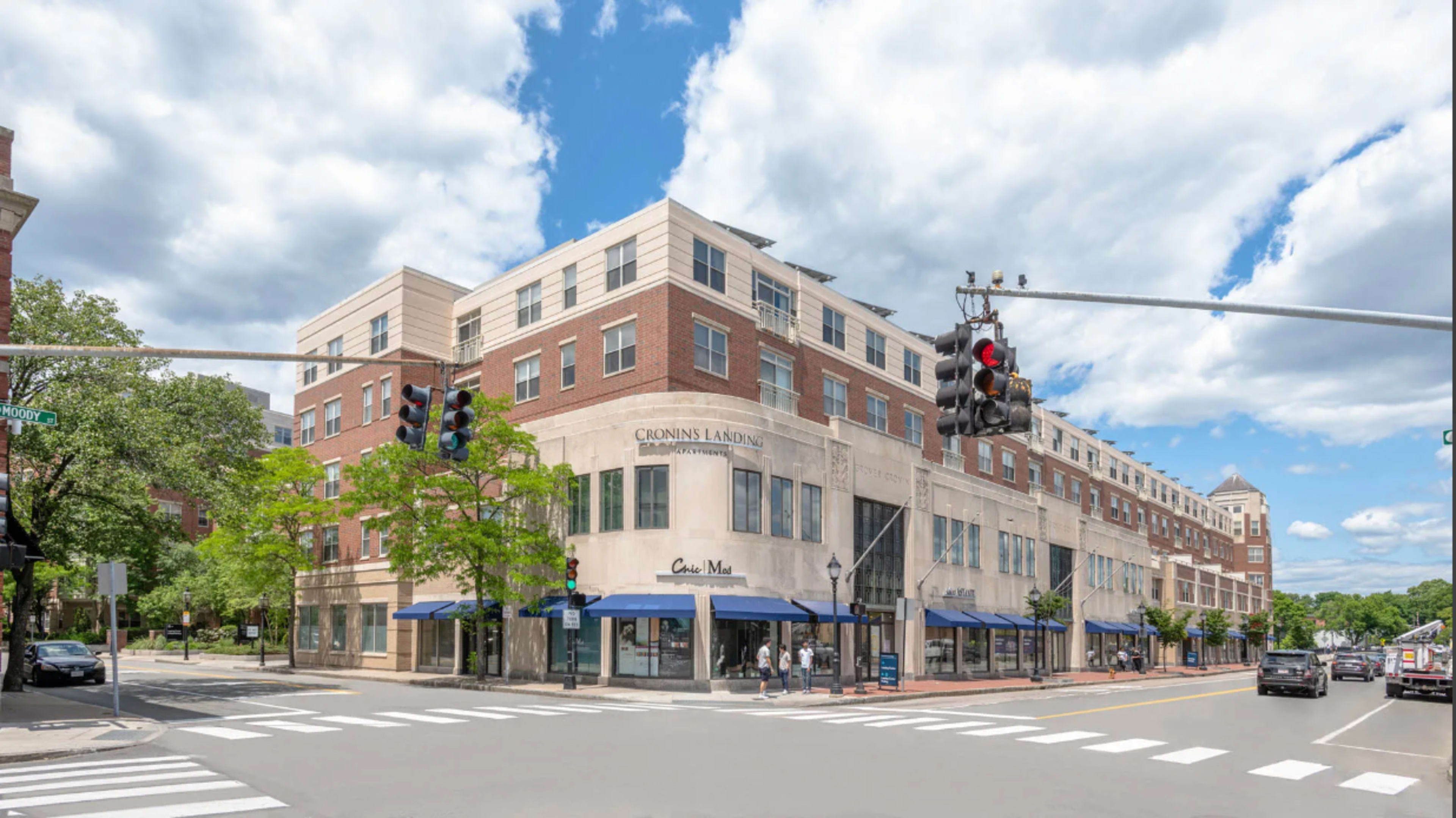 A multi-story building with retail storefronts and a traffic signal at a street intersection under a partly cloudy sky.