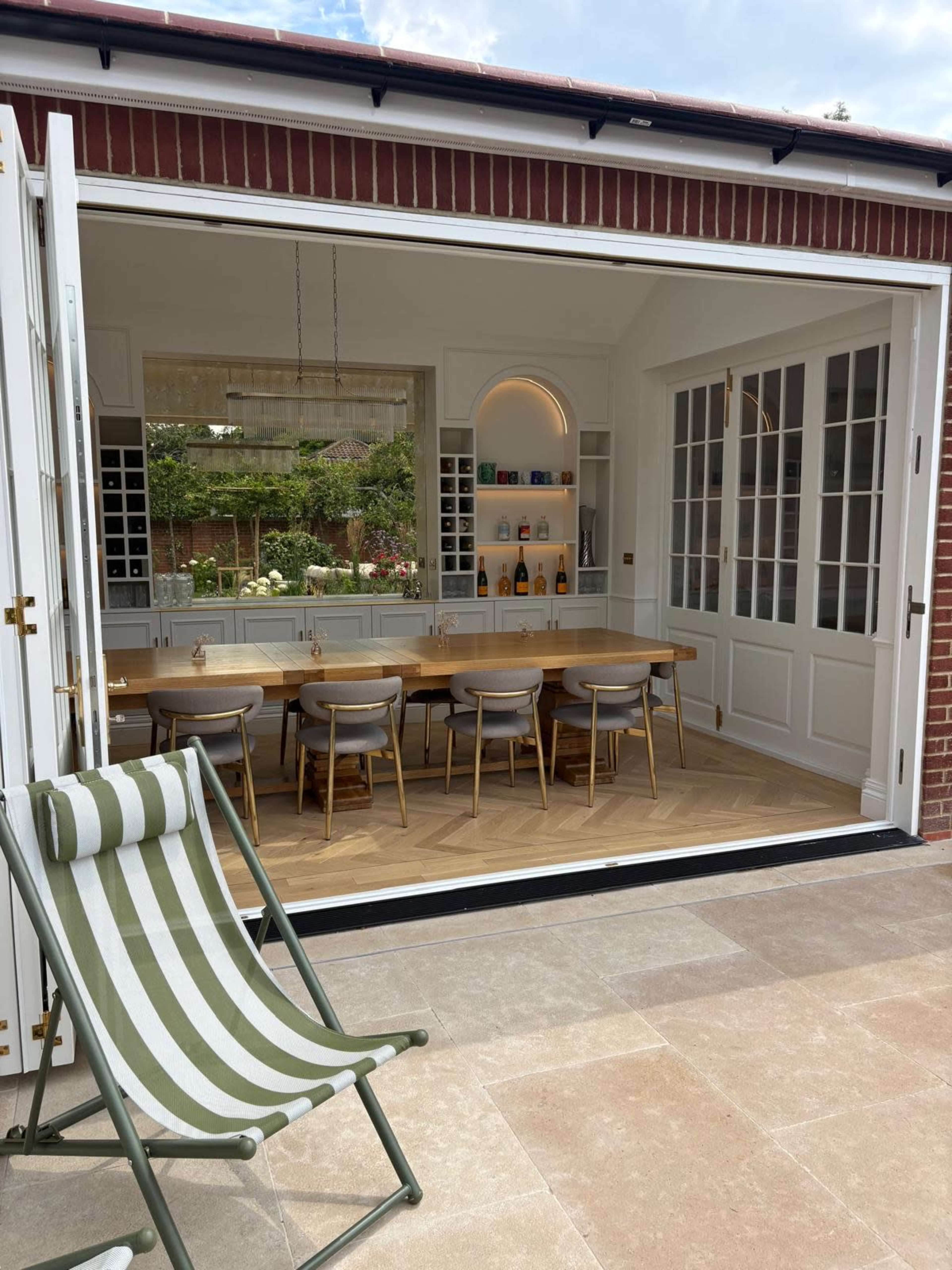 A dining area with a long wooden table and chairs is visible through an open sliding door, leading to a patio with a striped lounge chair.