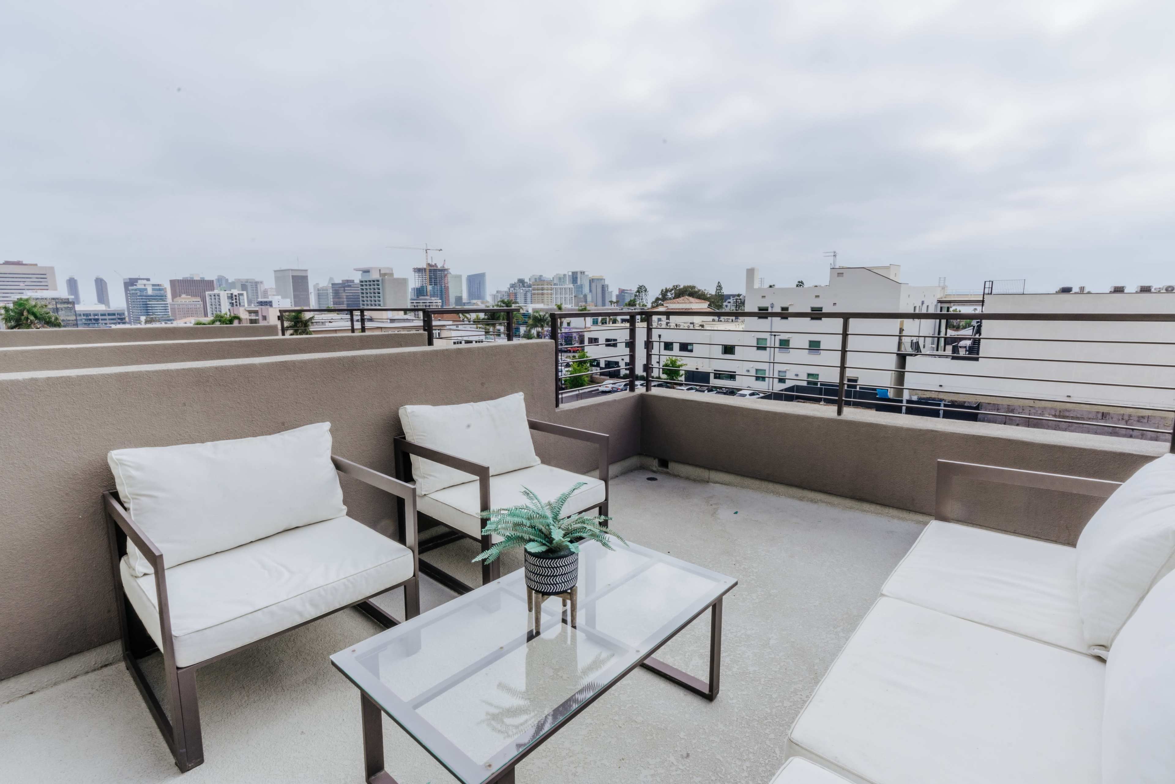 The image shows a rooftop terrace with white lounge furniture and a glass table, overlooking a city skyline under a gray sky.