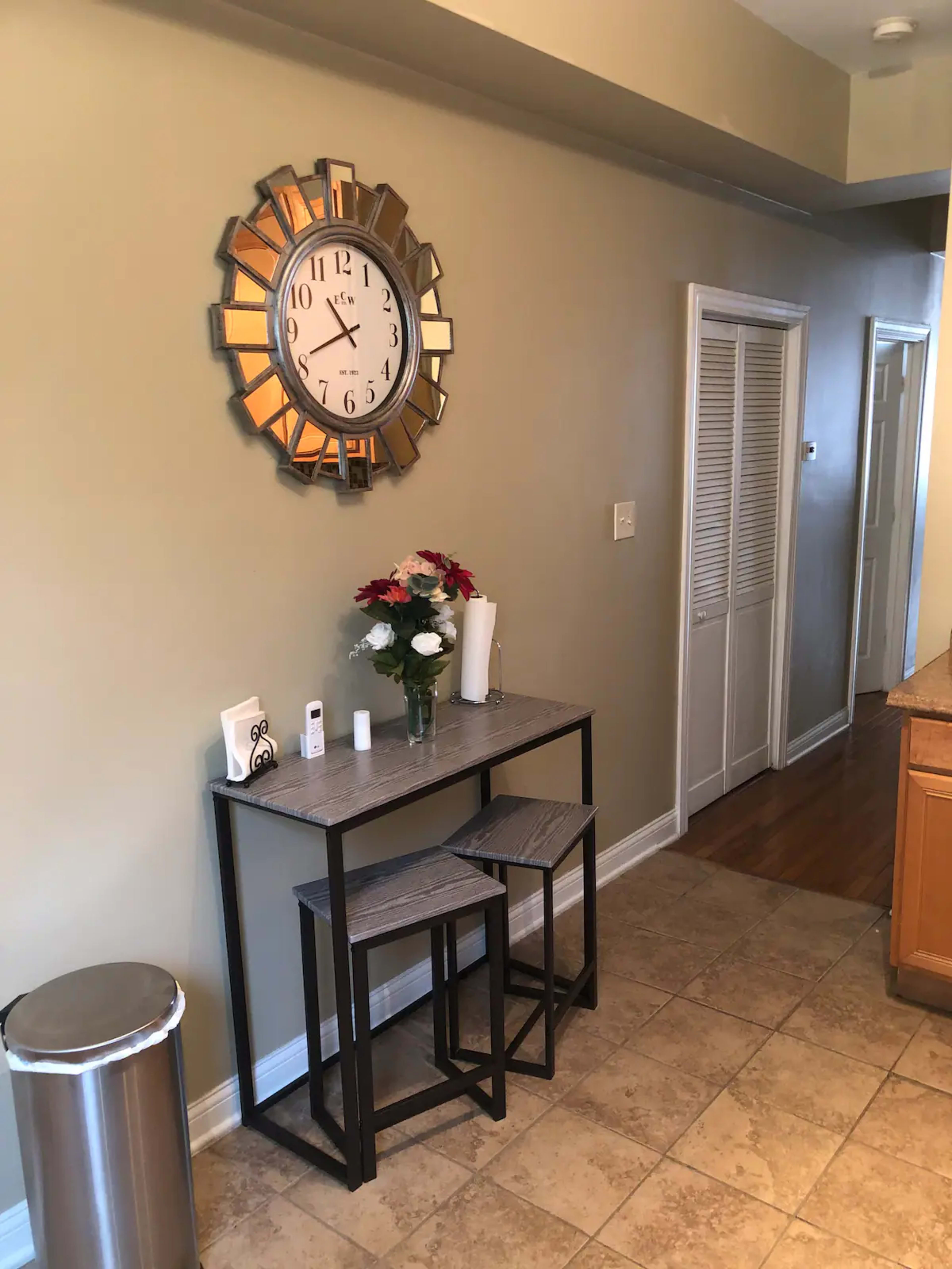 A narrow hallway with a table and two stools, a wall clock, a vase of flowers, and a waste bin.