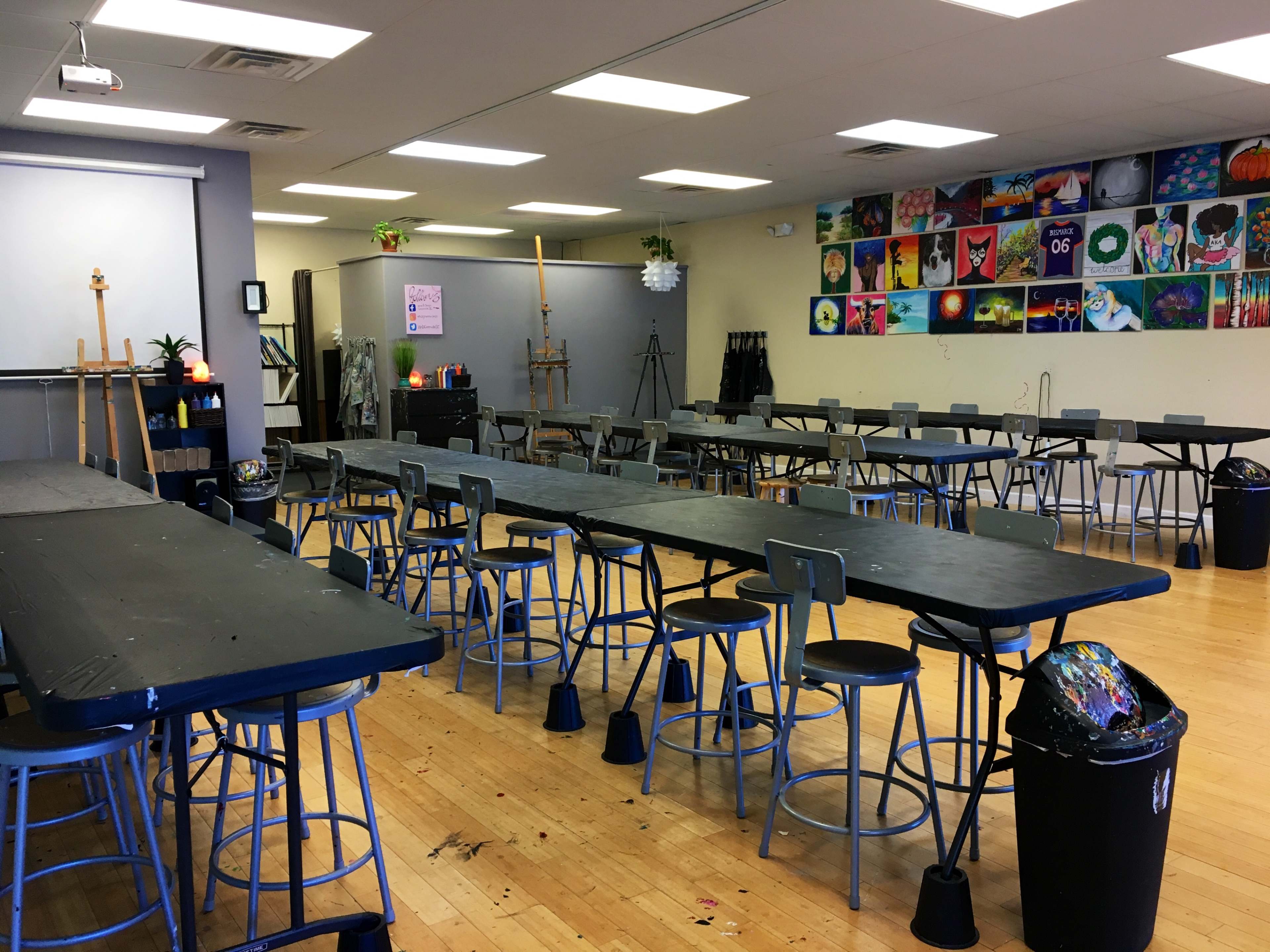 An art studio with multiple long tables and stools arranged in a spacious room, decorated with colorful paintings on the walls.