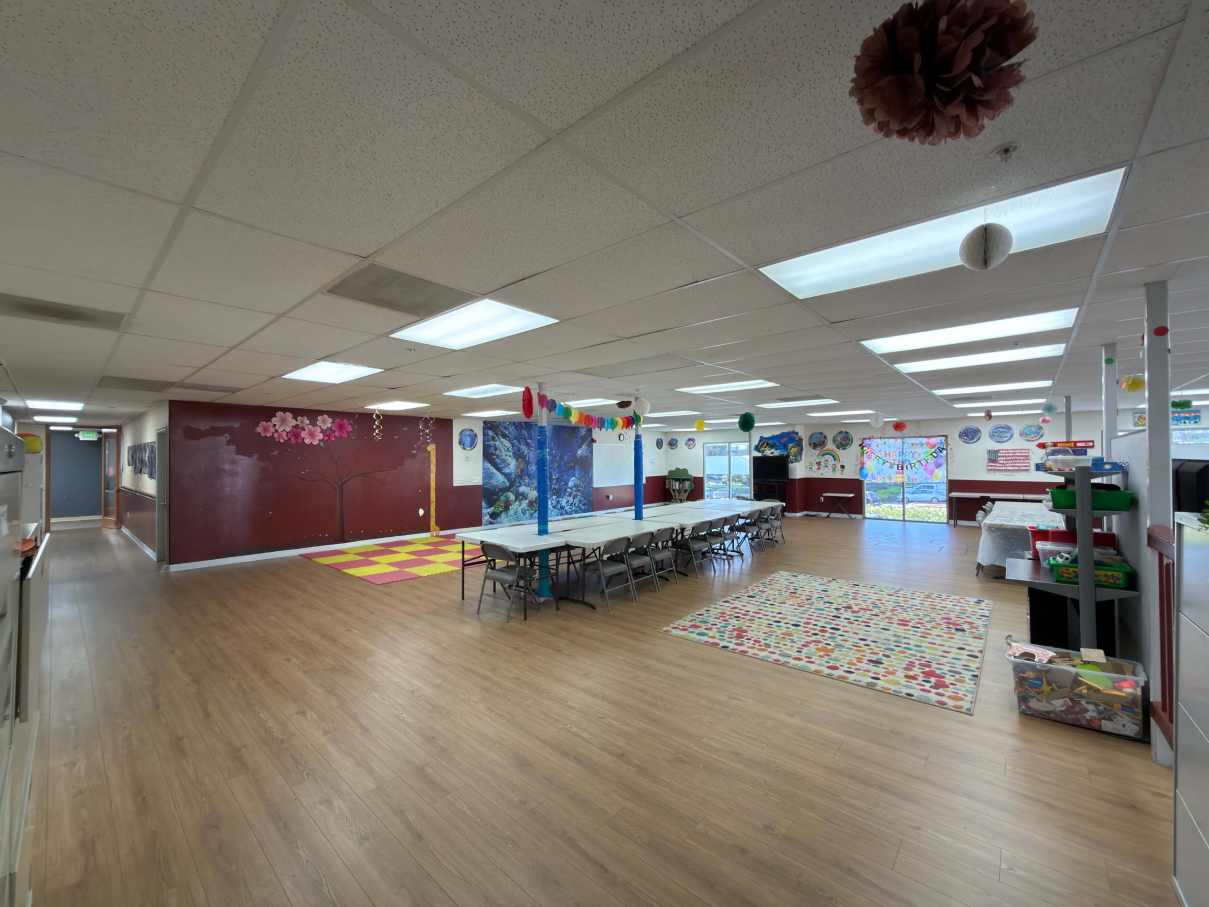 The image shows a spacious classroom with wooden flooring, several tables arranged in rows, colorful wall decorations, and various art supplies visible around the room.
