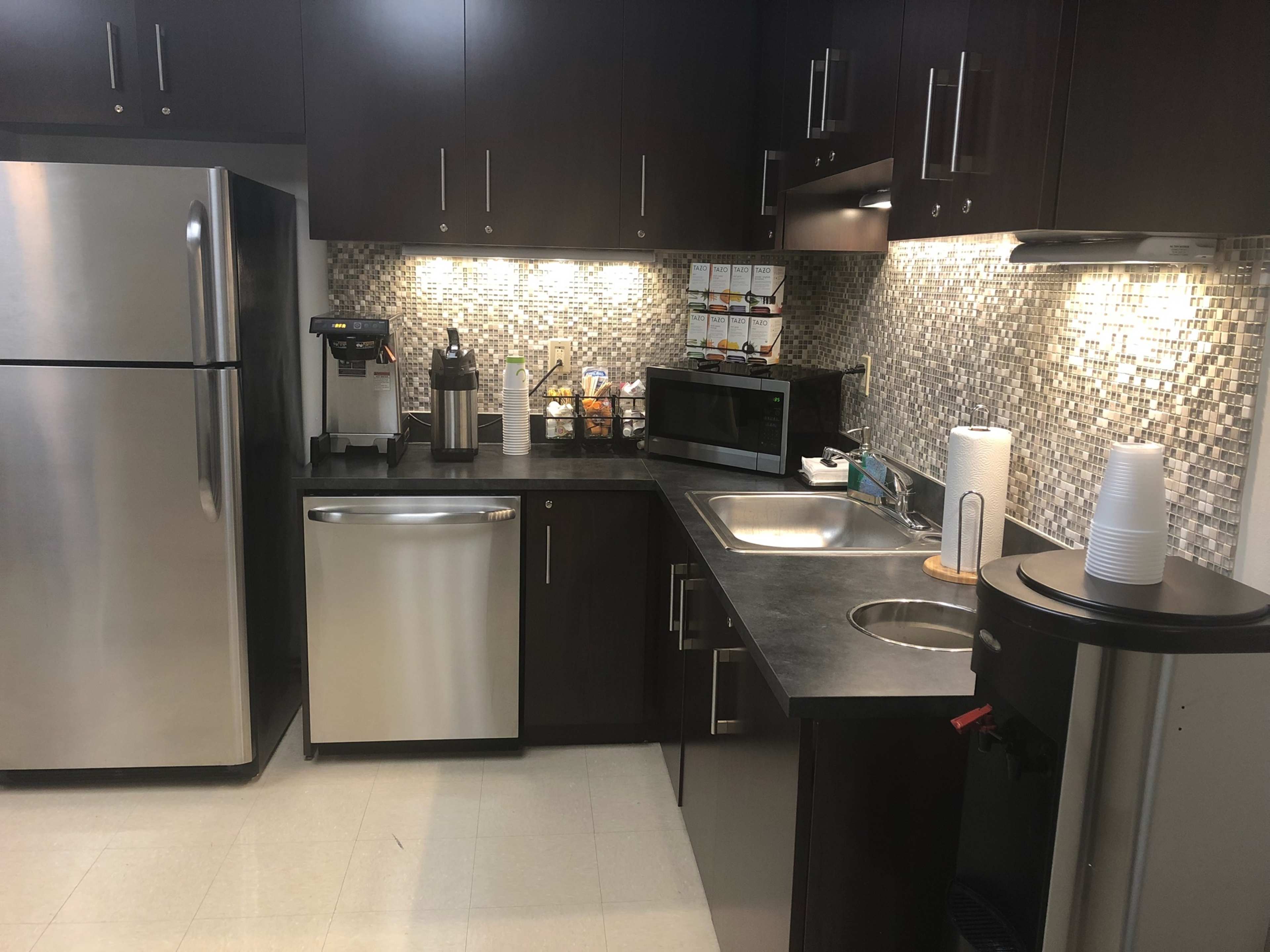 A modern kitchen with stainless steel appliances, including a refrigerator and dishwasher, a microwave, and a coffee machine, set against a tiled backsplash.