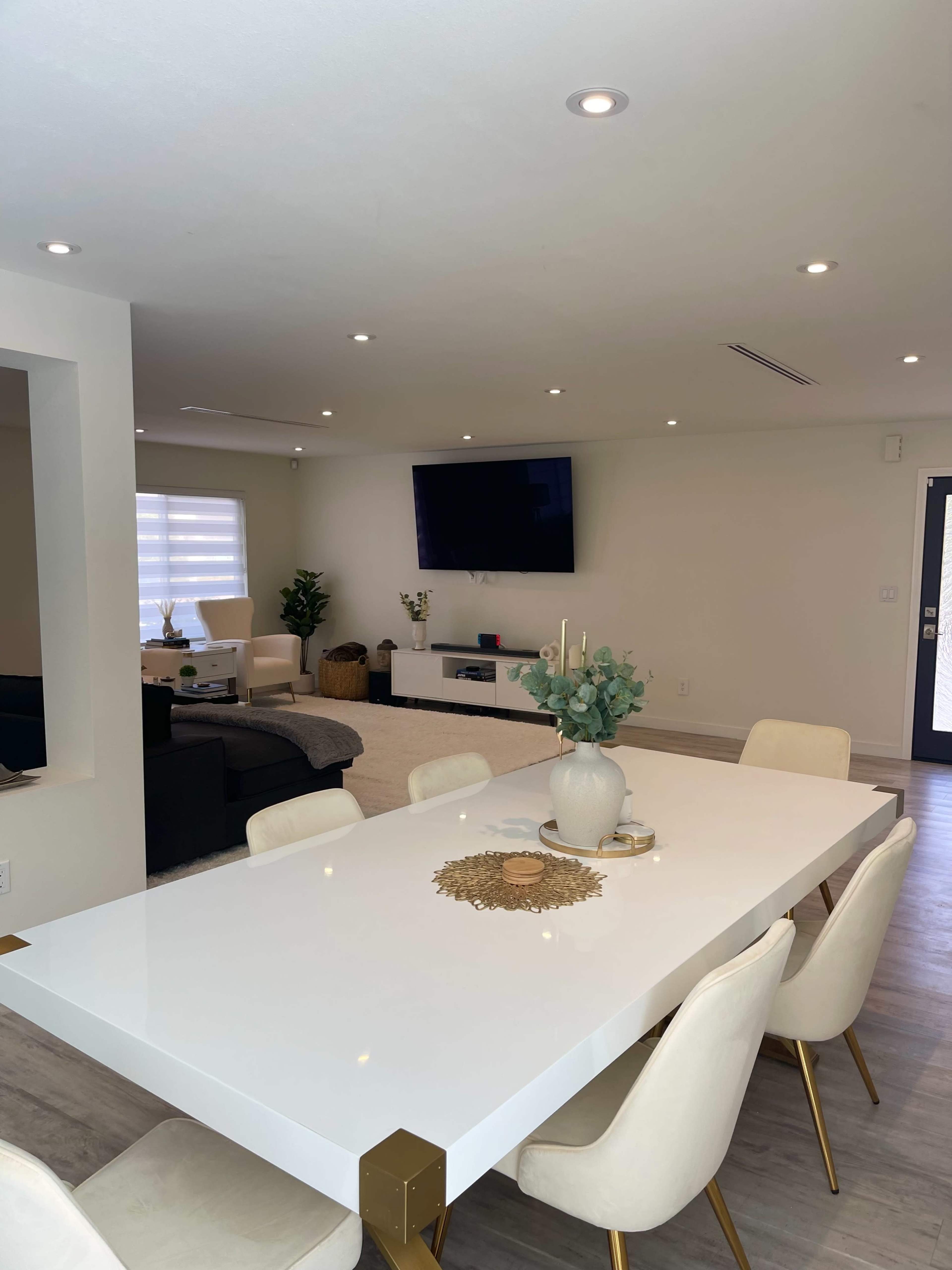 A modern living and dining area features a white table with beige chairs, a large television on the wall, and a minimalist decor style.