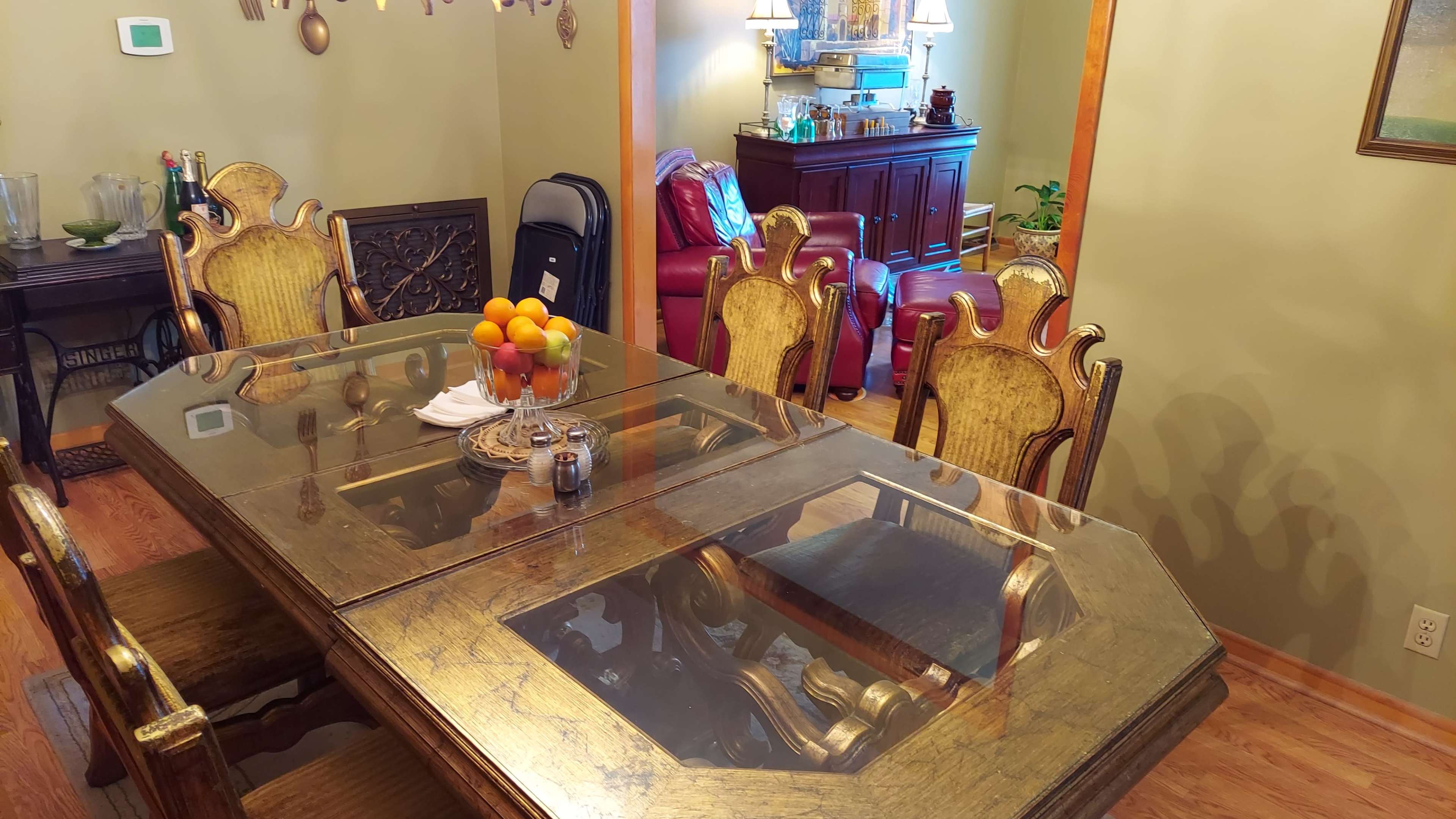 A dining table with a glass top is set with a bowl of oranges, surrounded by ornate wooden chairs in a room featuring various furnishings and decorations.
