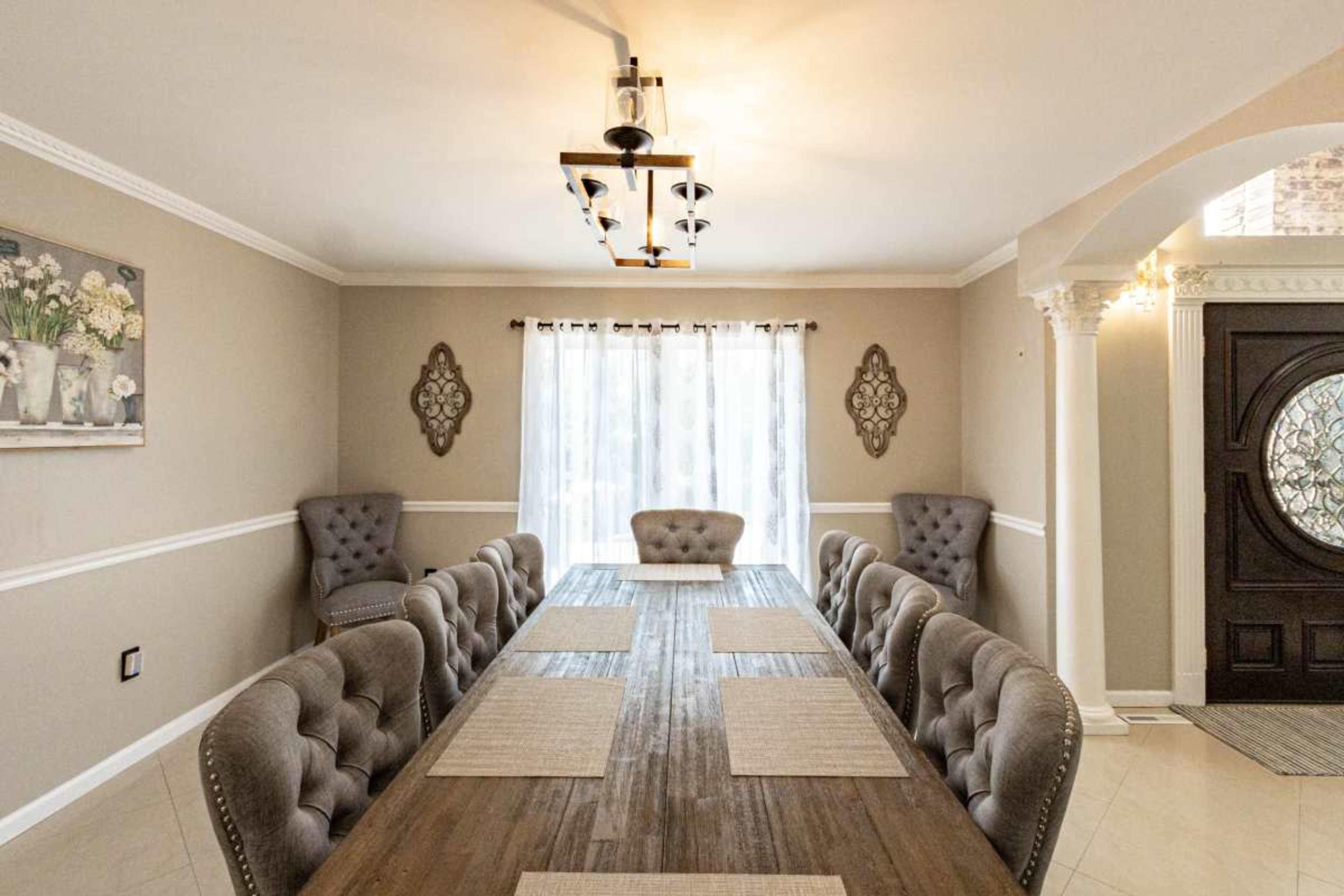 The image shows a spacious dining room with a long wooden table surrounded by upholstered chairs, featuring a large window with sheer curtains and decorative wall accents.