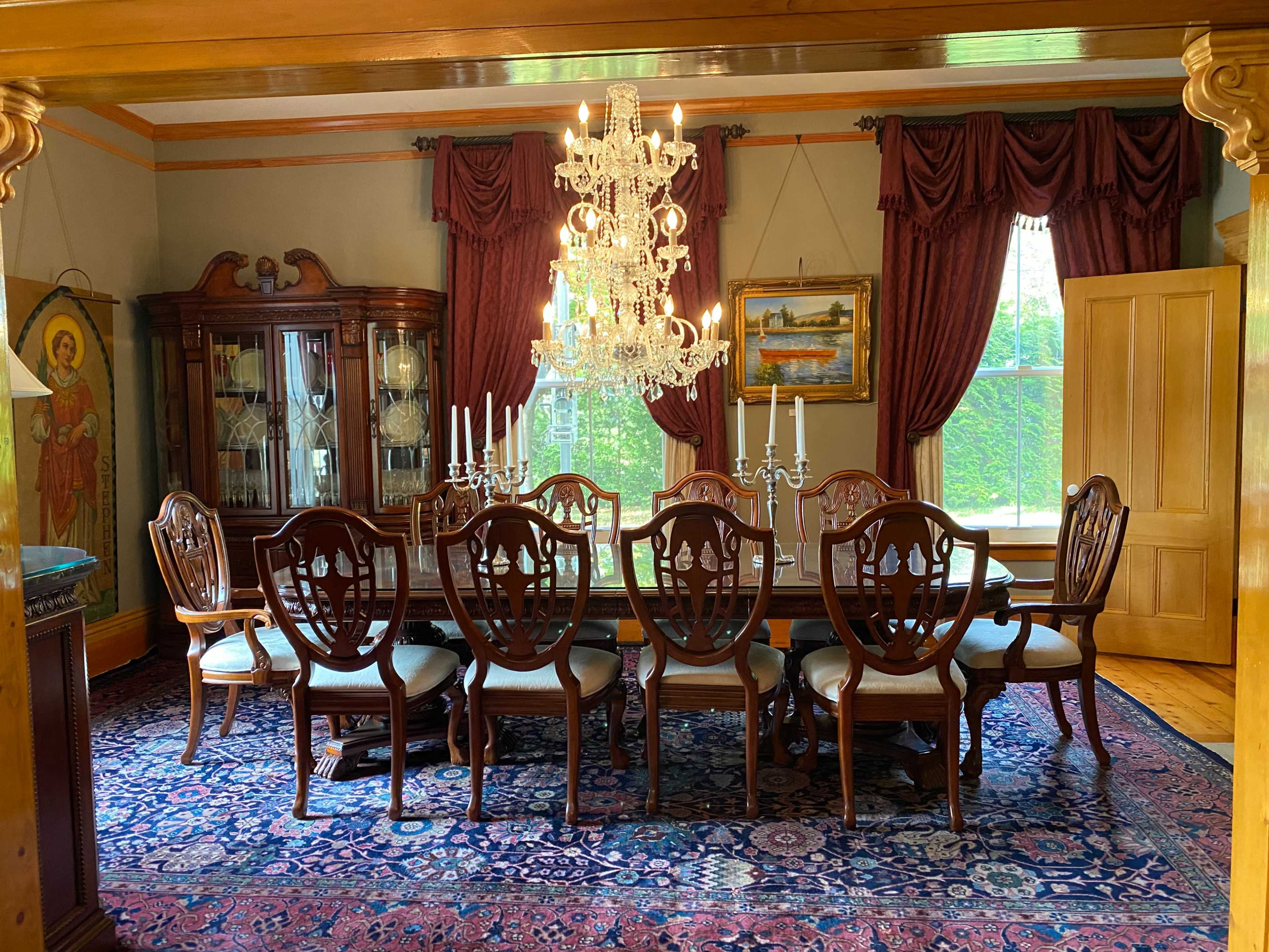 A formal dining room features a long wooden table surrounded by intricately designed chairs, a crystal chandelier above, and a display cabinet with decorative items.