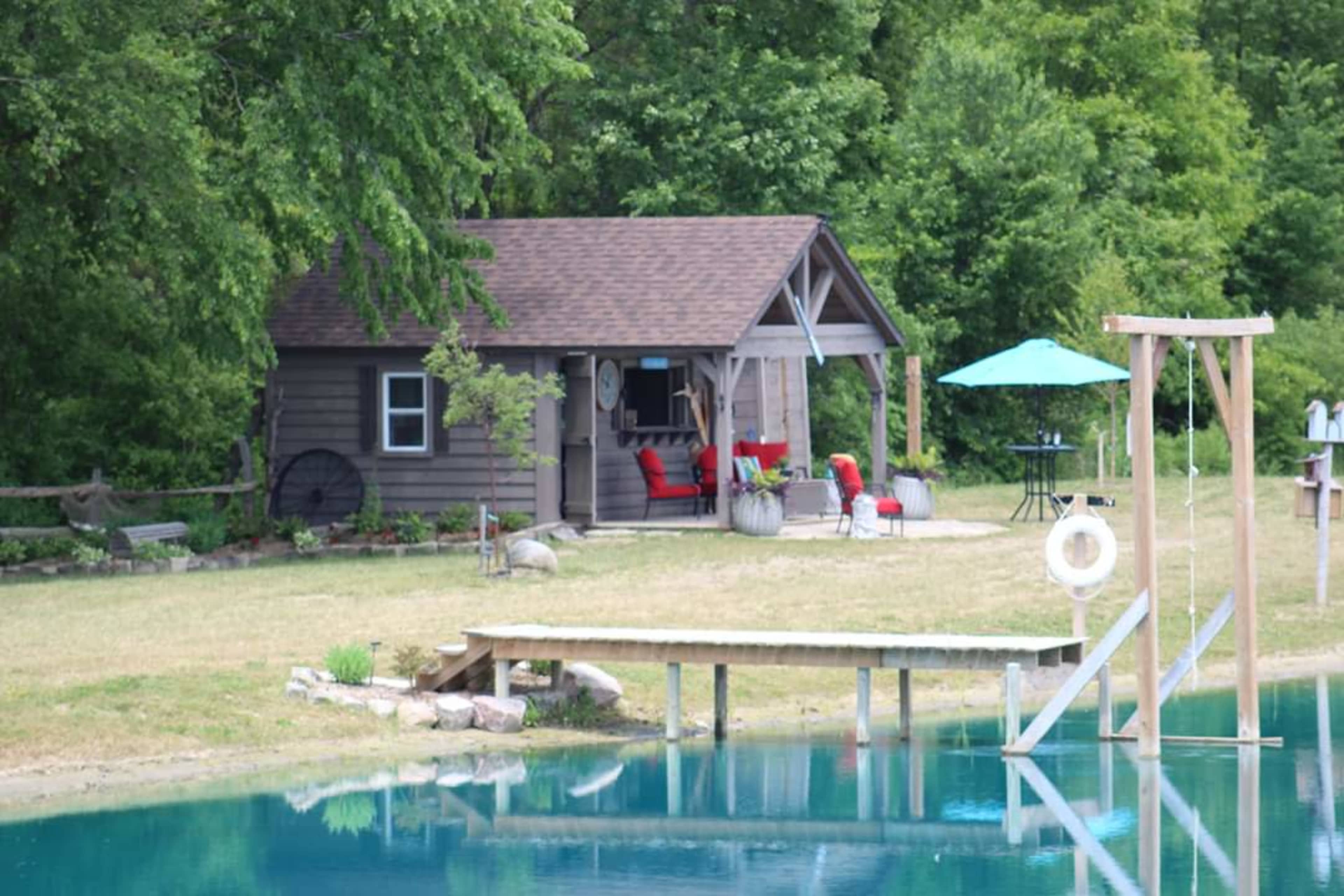 A small wooden cabin is situated beside a clear pond, with a patio featuring red chairs and a green umbrella.