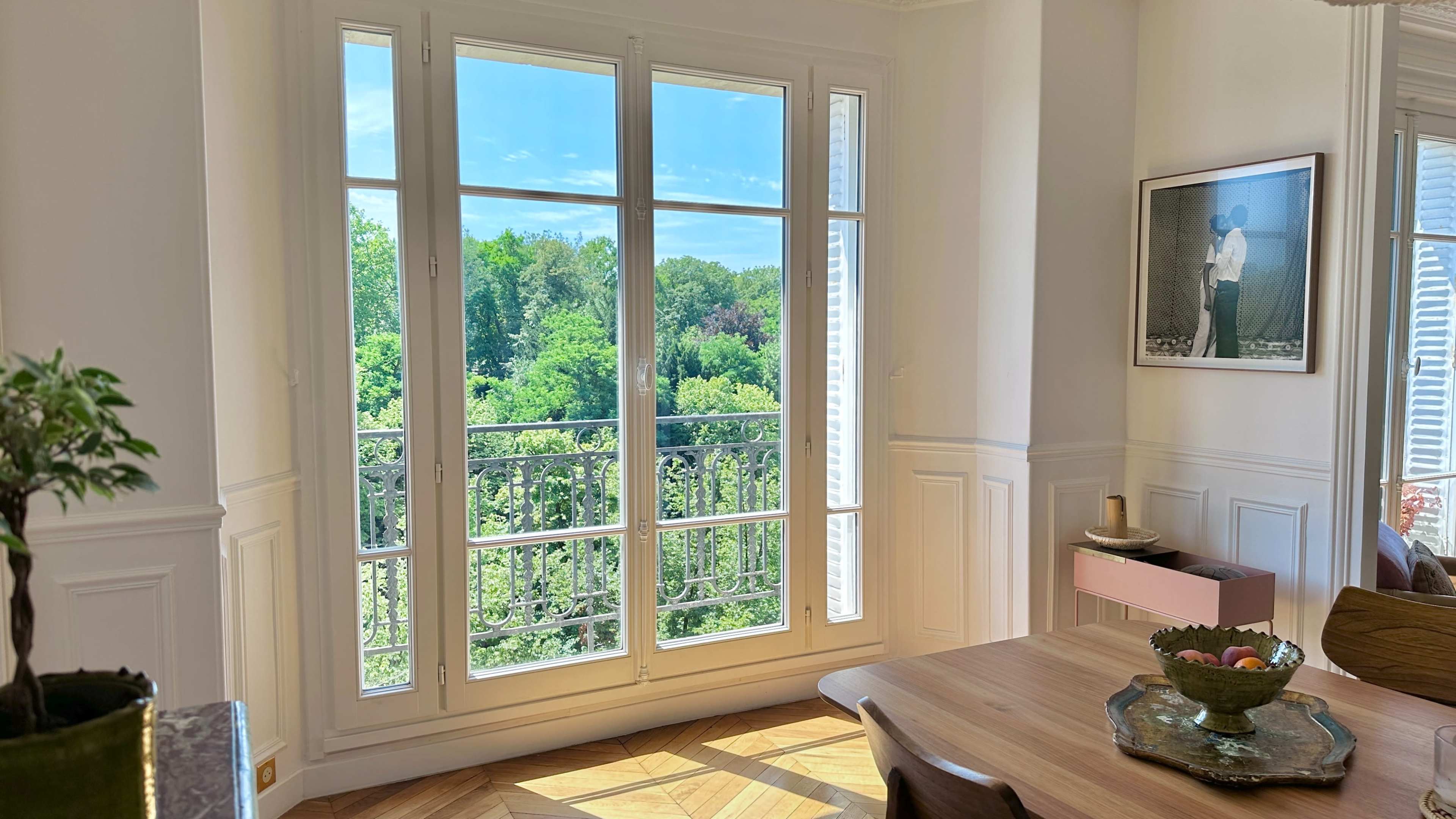 A bright room features a large window opening to a view of lush greenery outside, with a dining table set nearby.