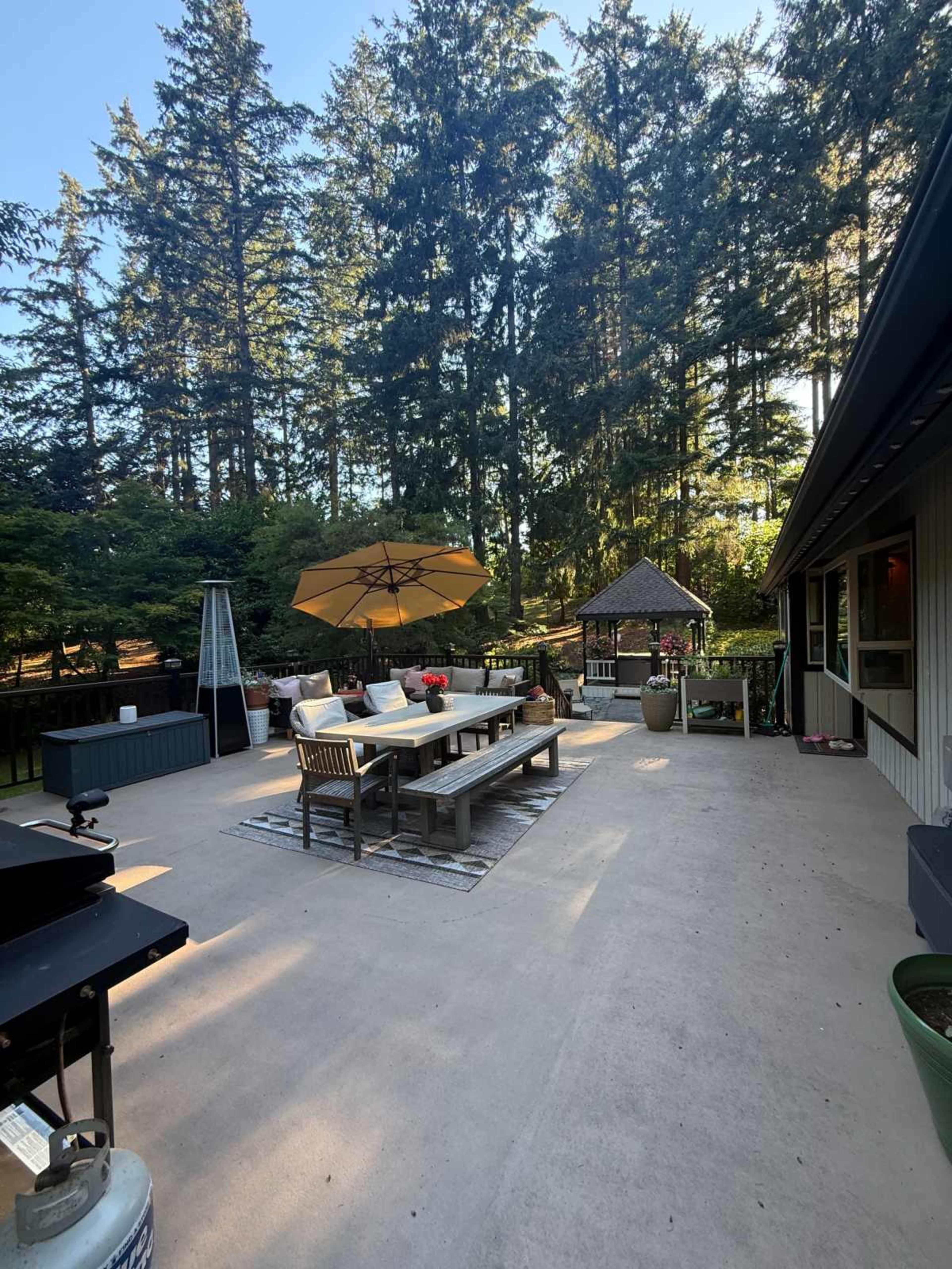 The image shows a patio area with outdoor furniture, including a long table, chairs, and an umbrella, surrounded by tall trees.