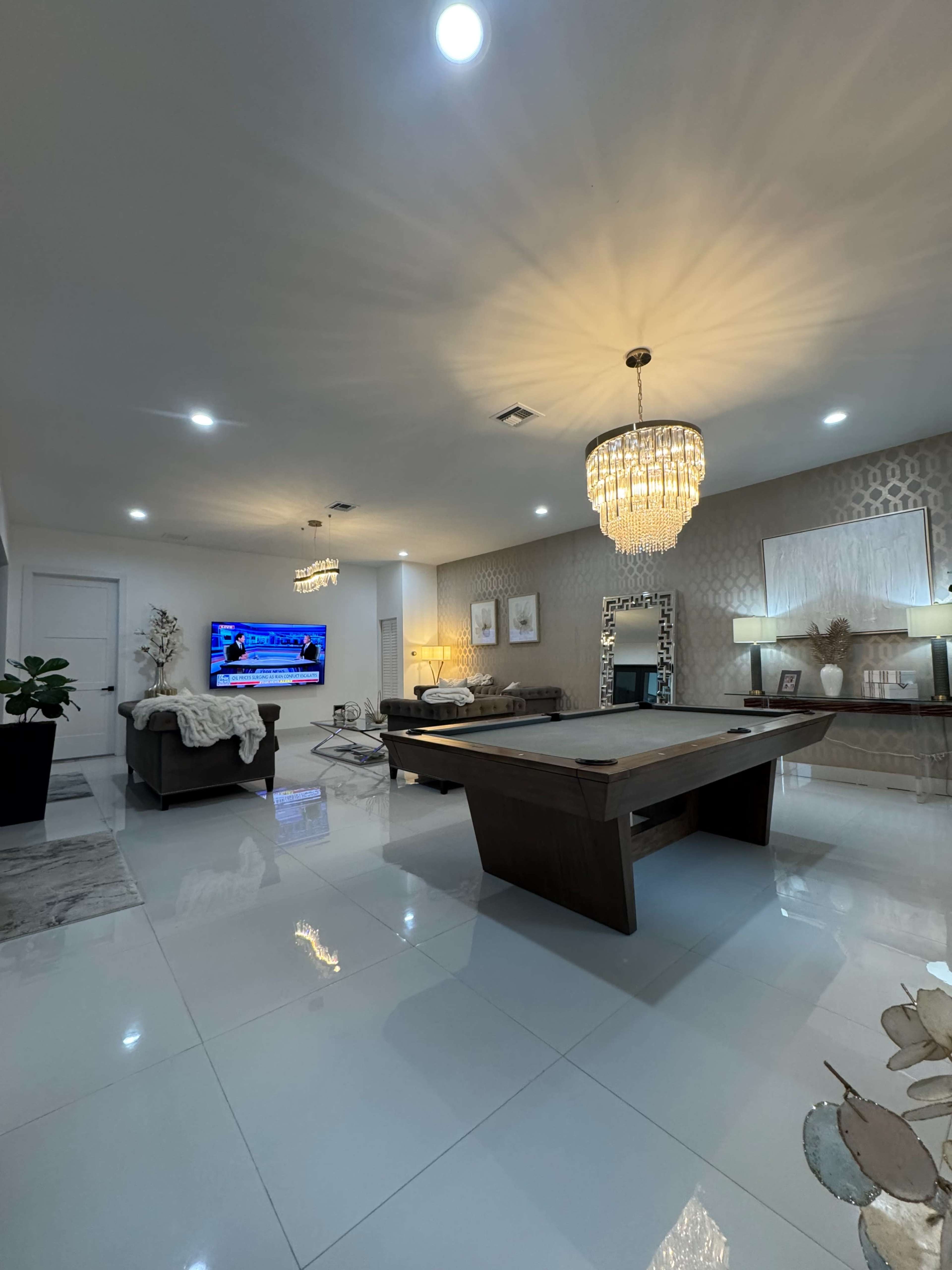 The image shows a modern living room featuring a pool table, a television mounted on the wall, and elegant chandeliers illuminating the space.
