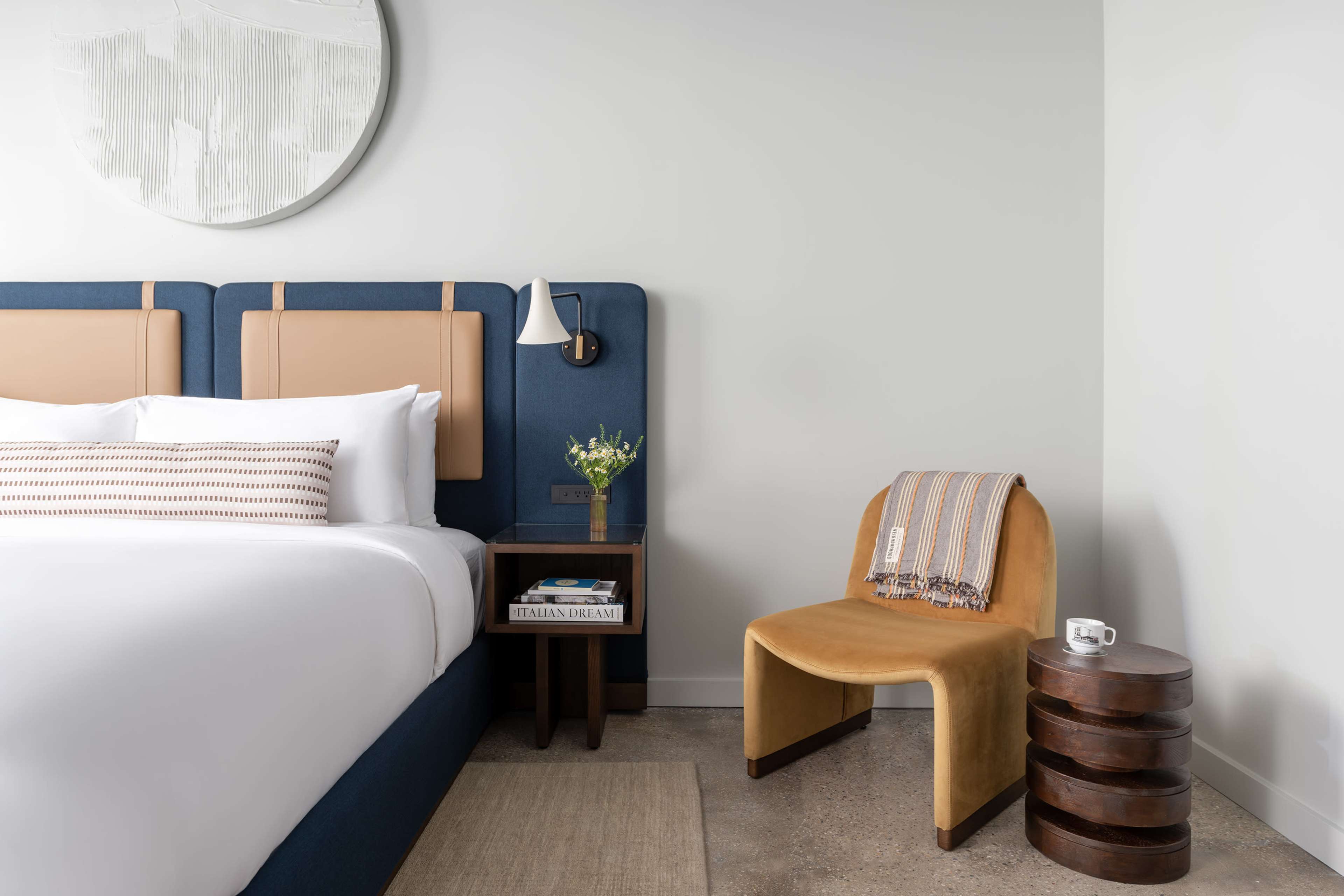 A neatly arranged hotel room features a bed with a padded headboard, a small wooden bedside table, and a modern chair beside it.
