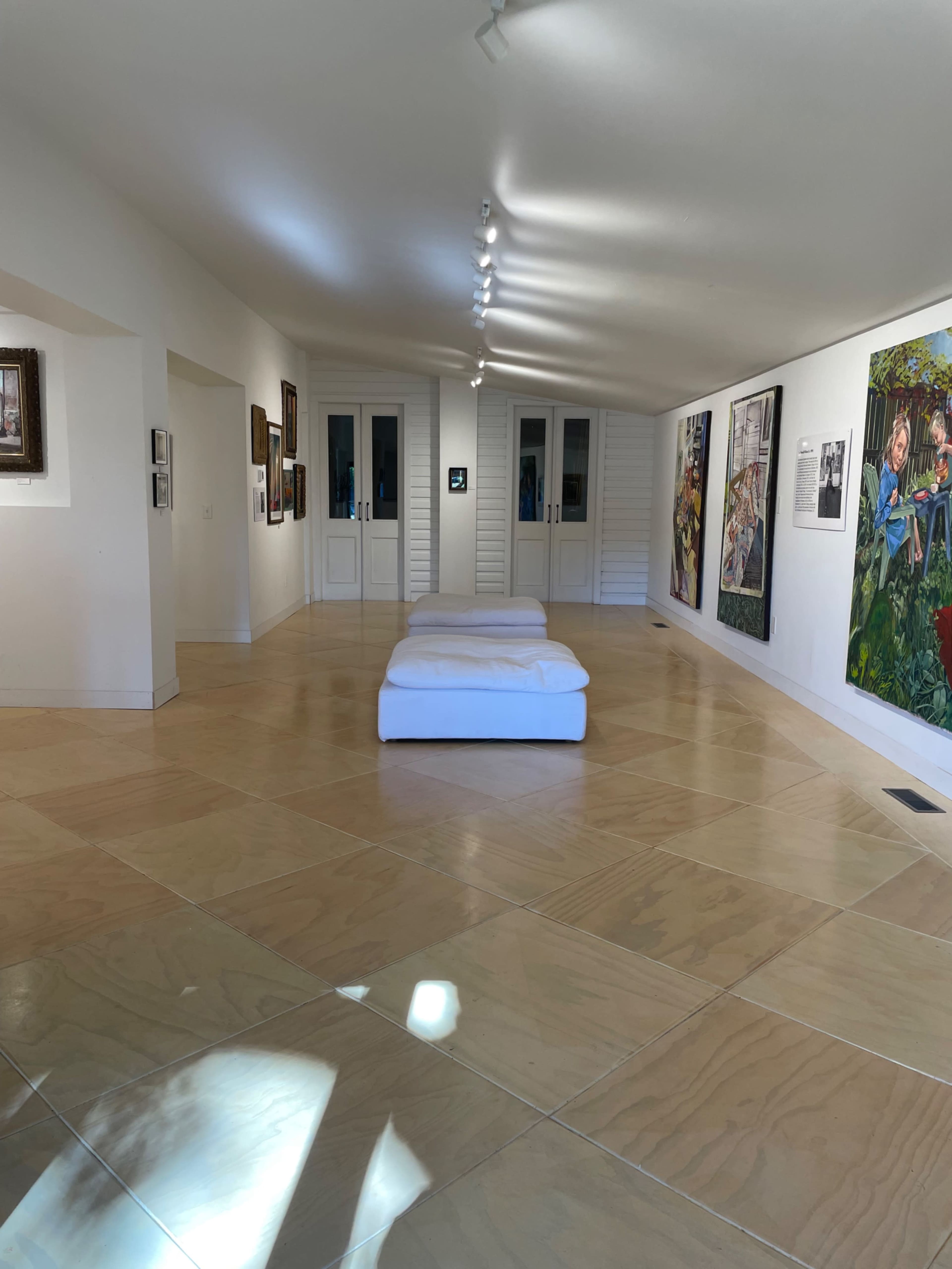 The image shows a minimalist art gallery with white walls, several pieces of art displayed, and three white mattresses arranged on the polished floor.