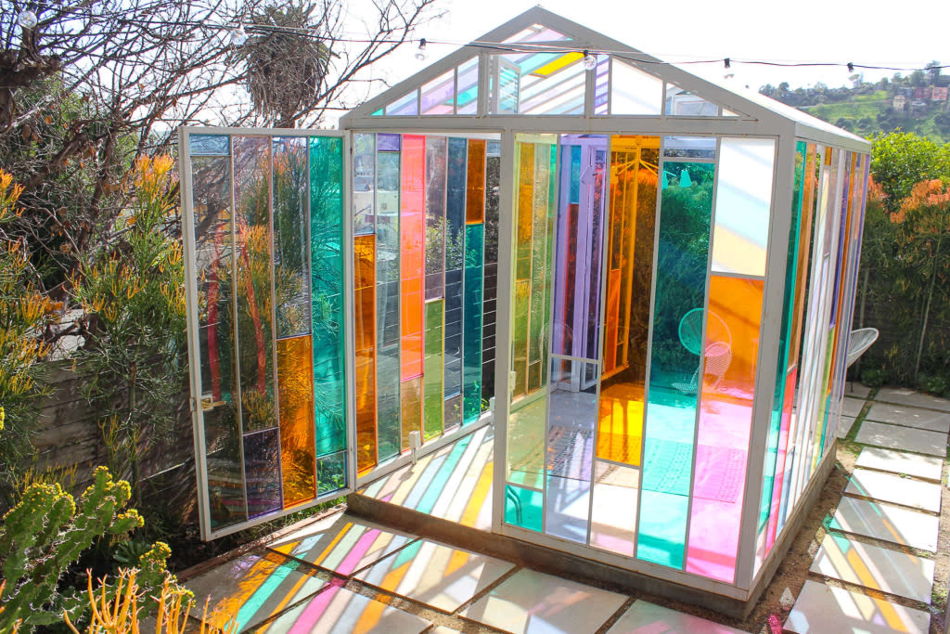 A colorful glass greenhouse with multicolored panels and an open doorway, situated in a garden with plants and outdoor tiles.