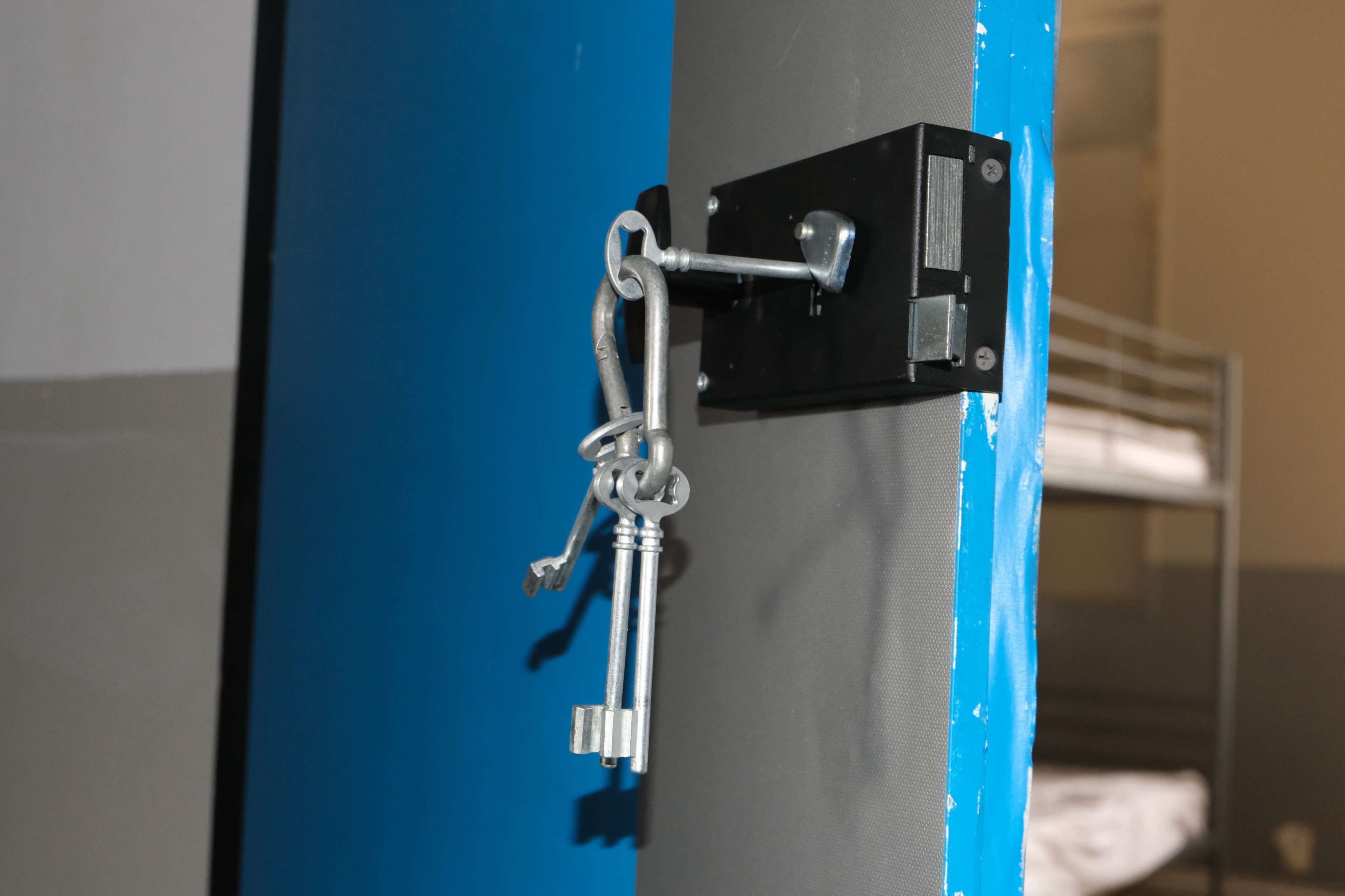 A set of metal keys hangs on a black lock attached to a blue door, with a bed frame visible in the background.