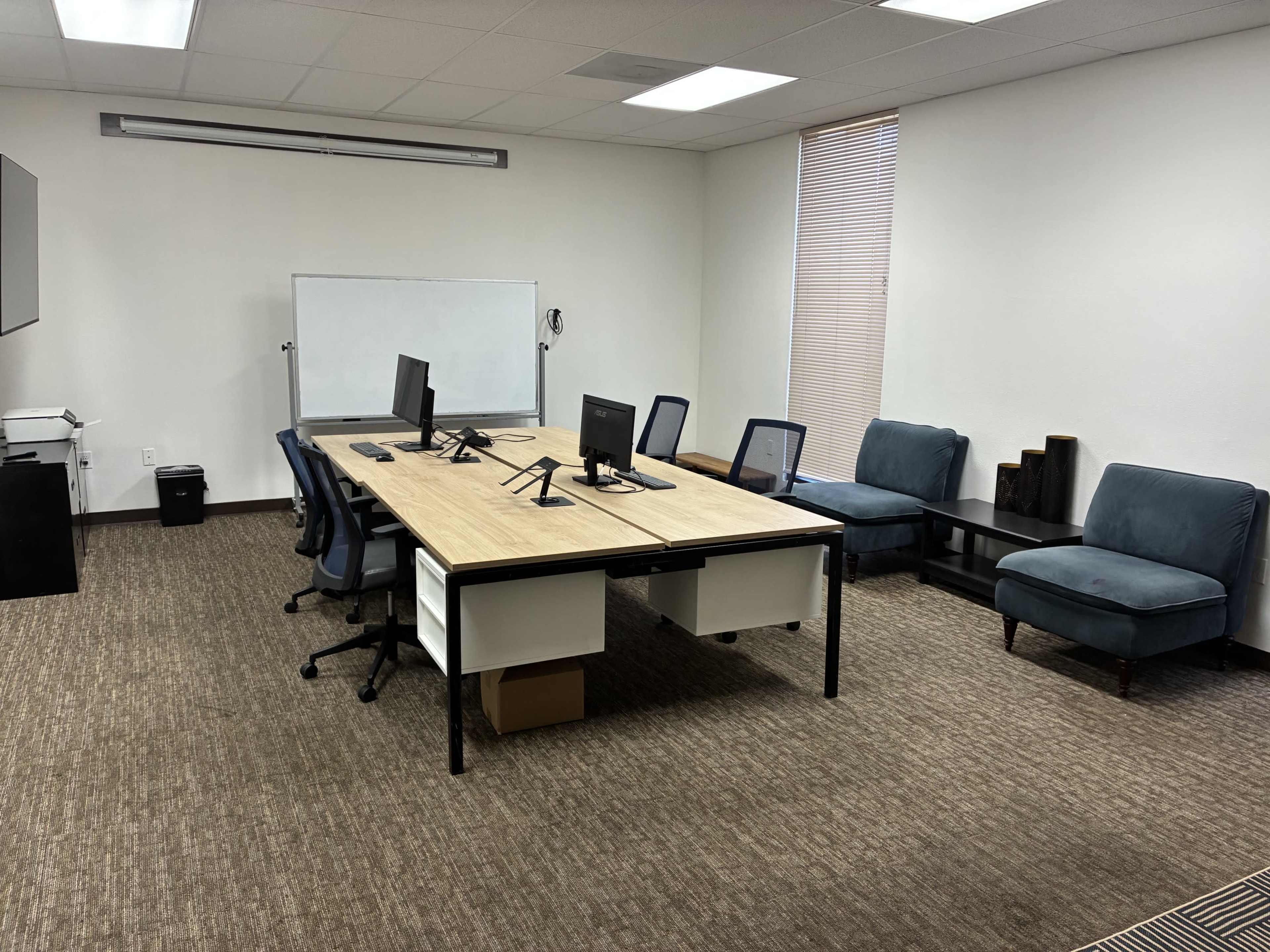 The image shows an office space with a large table, two computer monitors, six chairs, a whiteboard, and two blue chairs along the wall.
