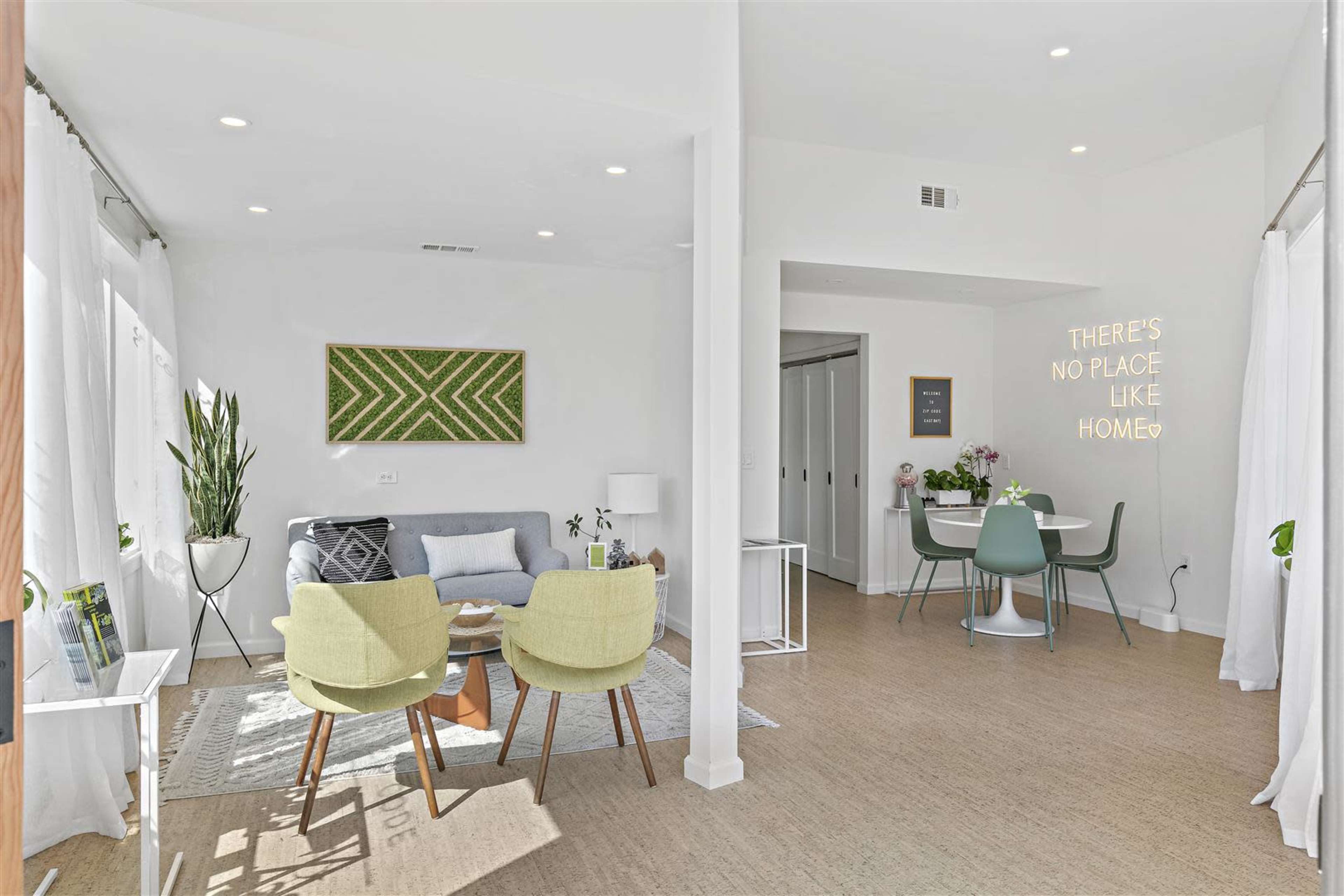 A bright, modern living space featuring a light gray sofa, two green chairs, a dining area with a small table, and decorative plants.