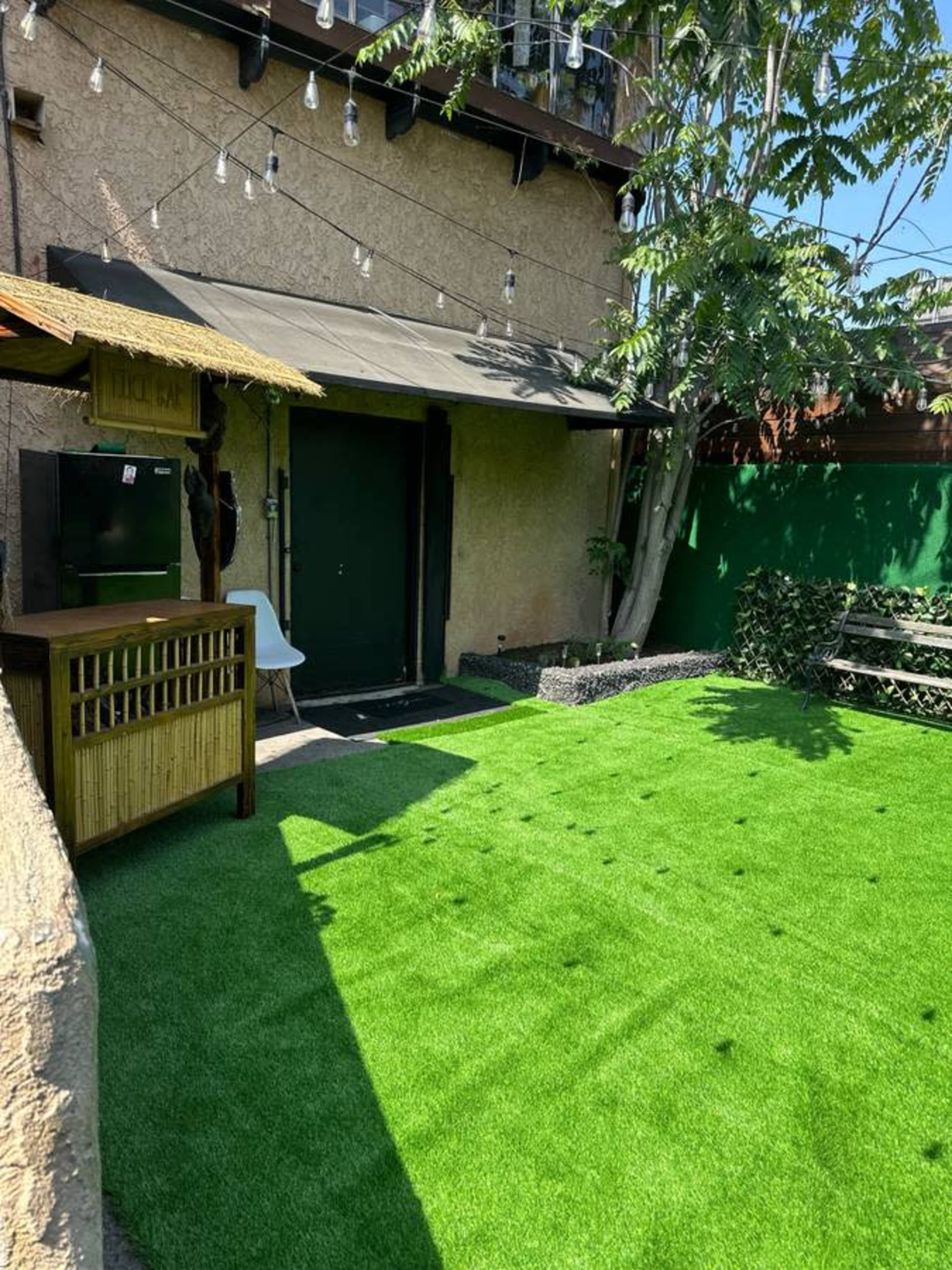 A small backyard features artificial grass, a wooden seating area, and a door leading to a house, surrounded by plants and decorative lights.