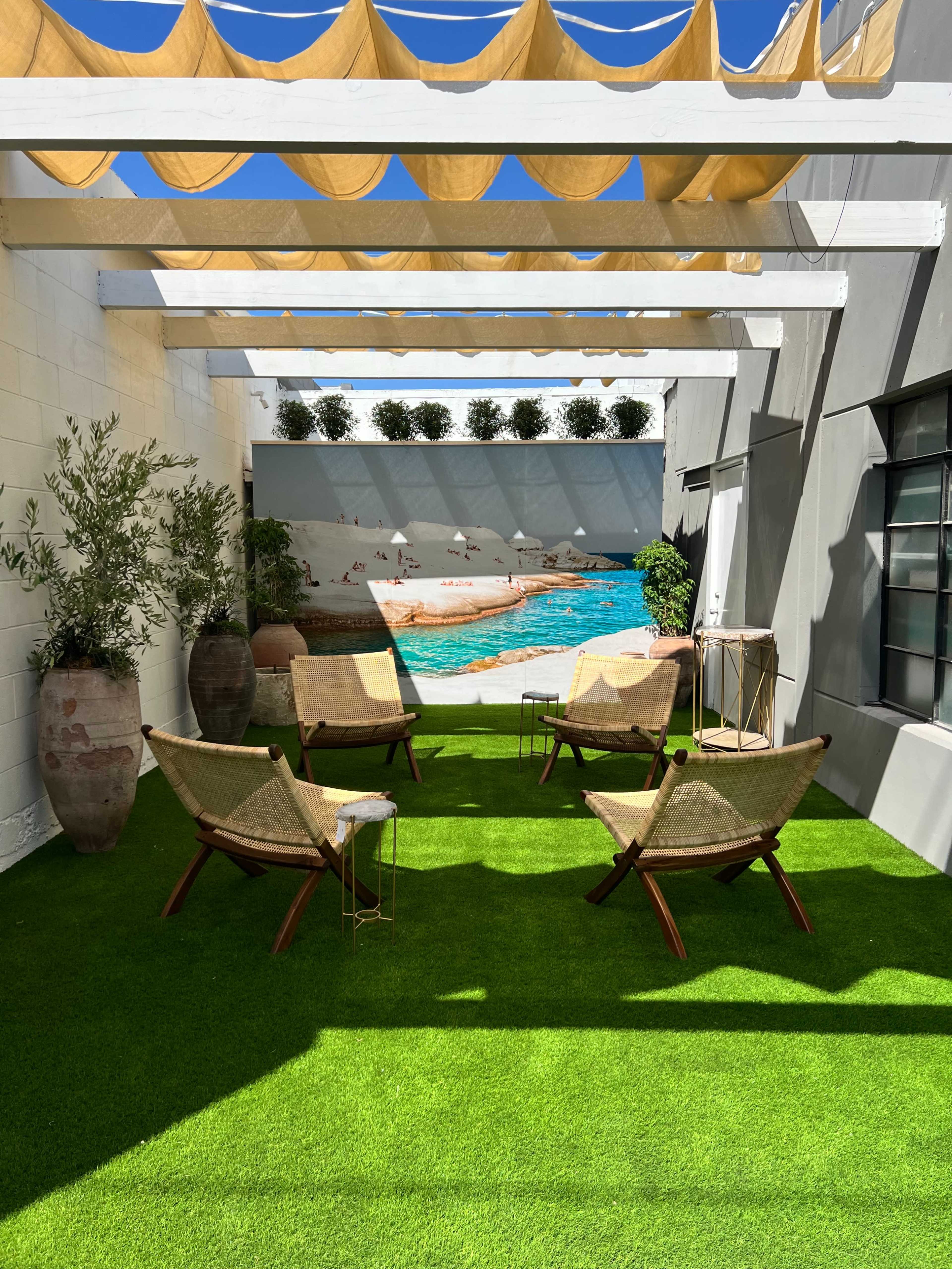 The image shows a sunlit outdoor patio with green artificial grass, four woven chairs, potted plants, and a wall mural depicting a pool scene.