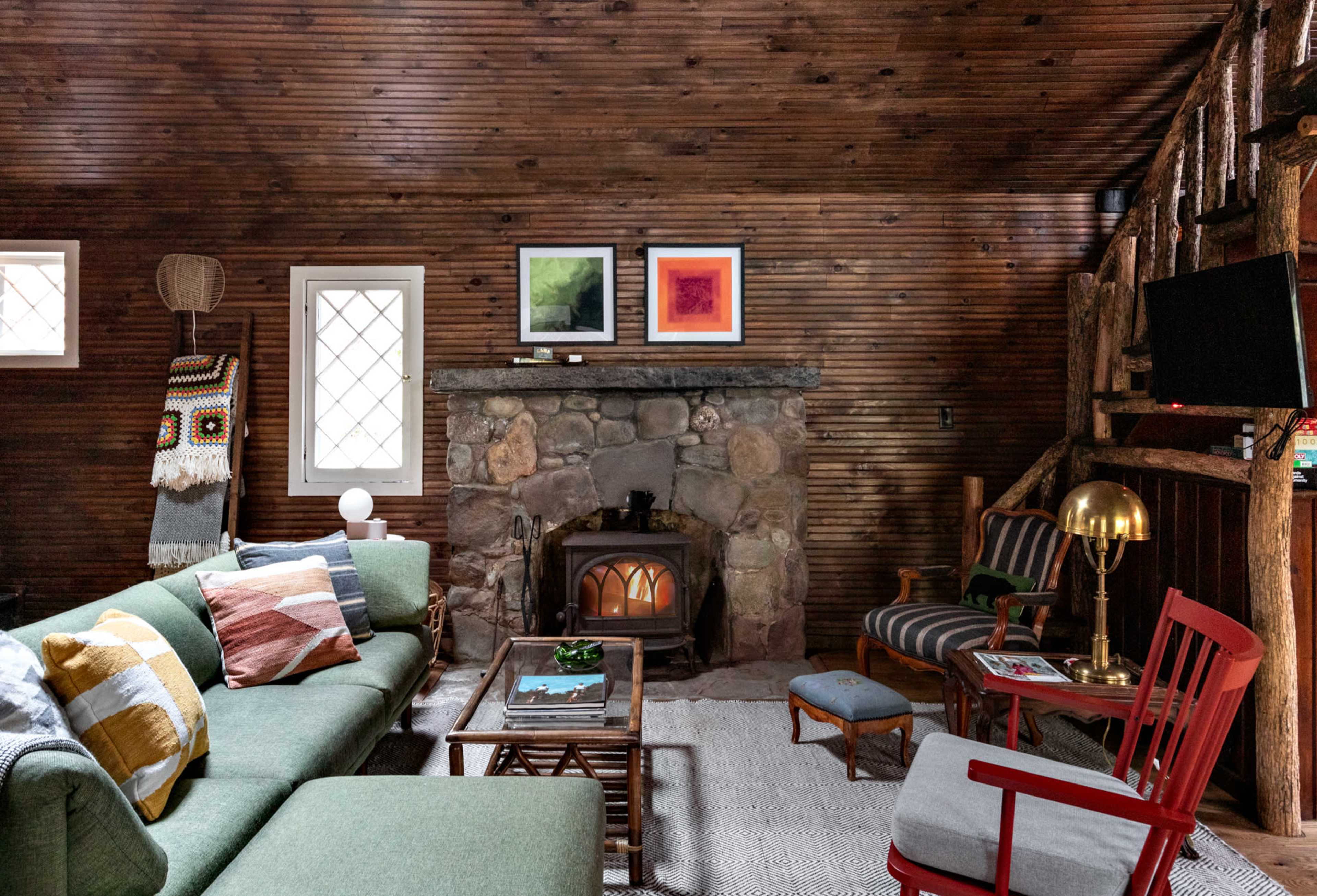The interior of a rustic cabin features a stone fireplace, wooden walls, and a mix of furniture including a green sofa, a wooden coffee table, and various seating arrangements.