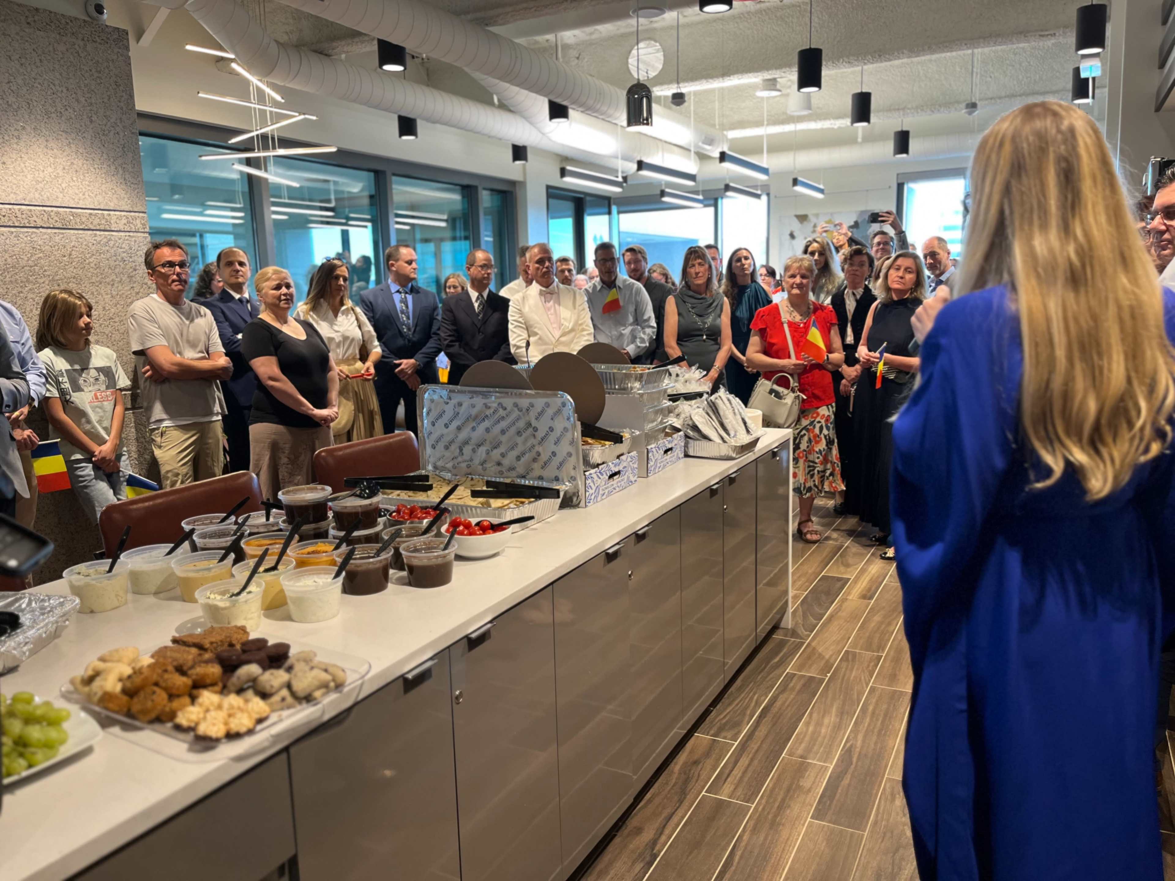 A large group of people gathers in a modern kitchen area, facing a speaker, with various food items displayed on a counter.