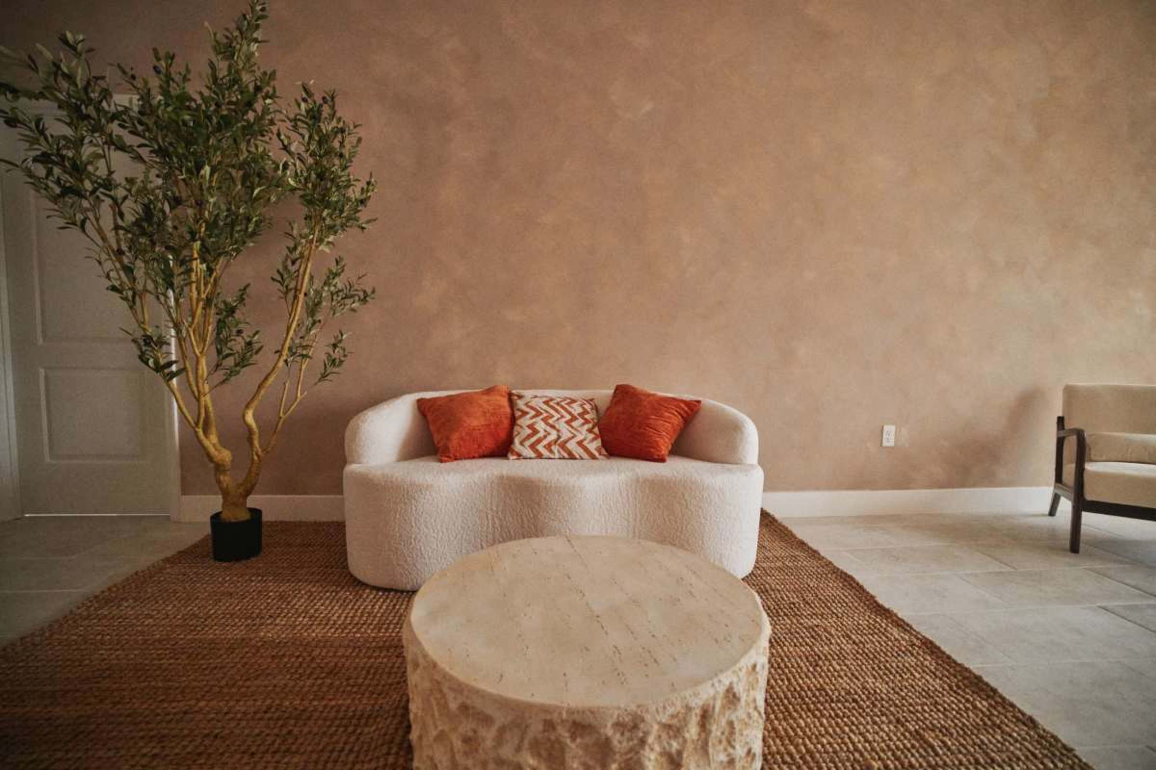 The scene features a cozy, minimalist living space with a white, curved sofa adorned with orange pillows, a round wooden coffee table, and a small potted tree, all set against a warm, textured wall.