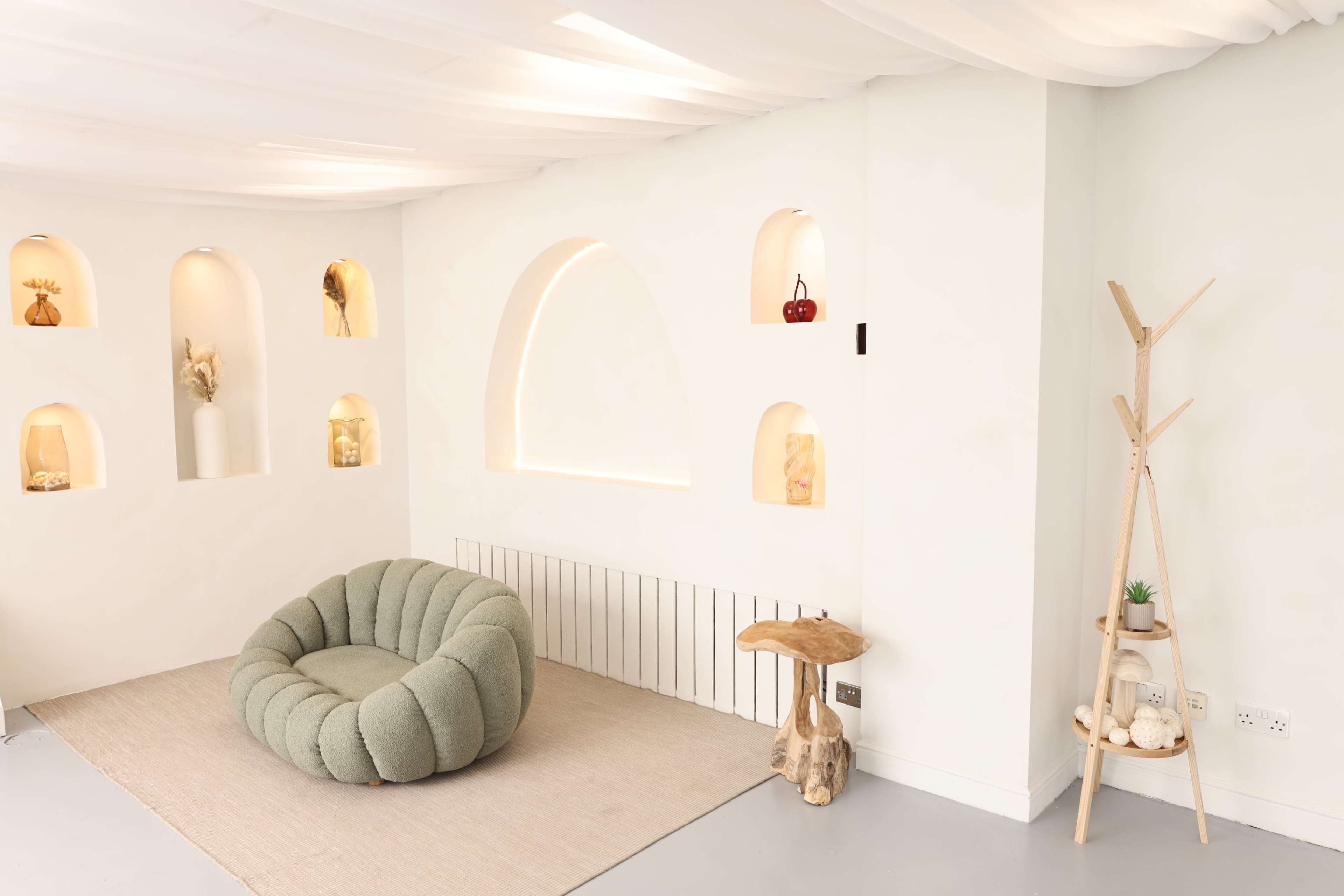 The image shows a modern interior space featuring a round green upholstered chair, decorative niches in a white wall, and a wooden stand with a small plant.