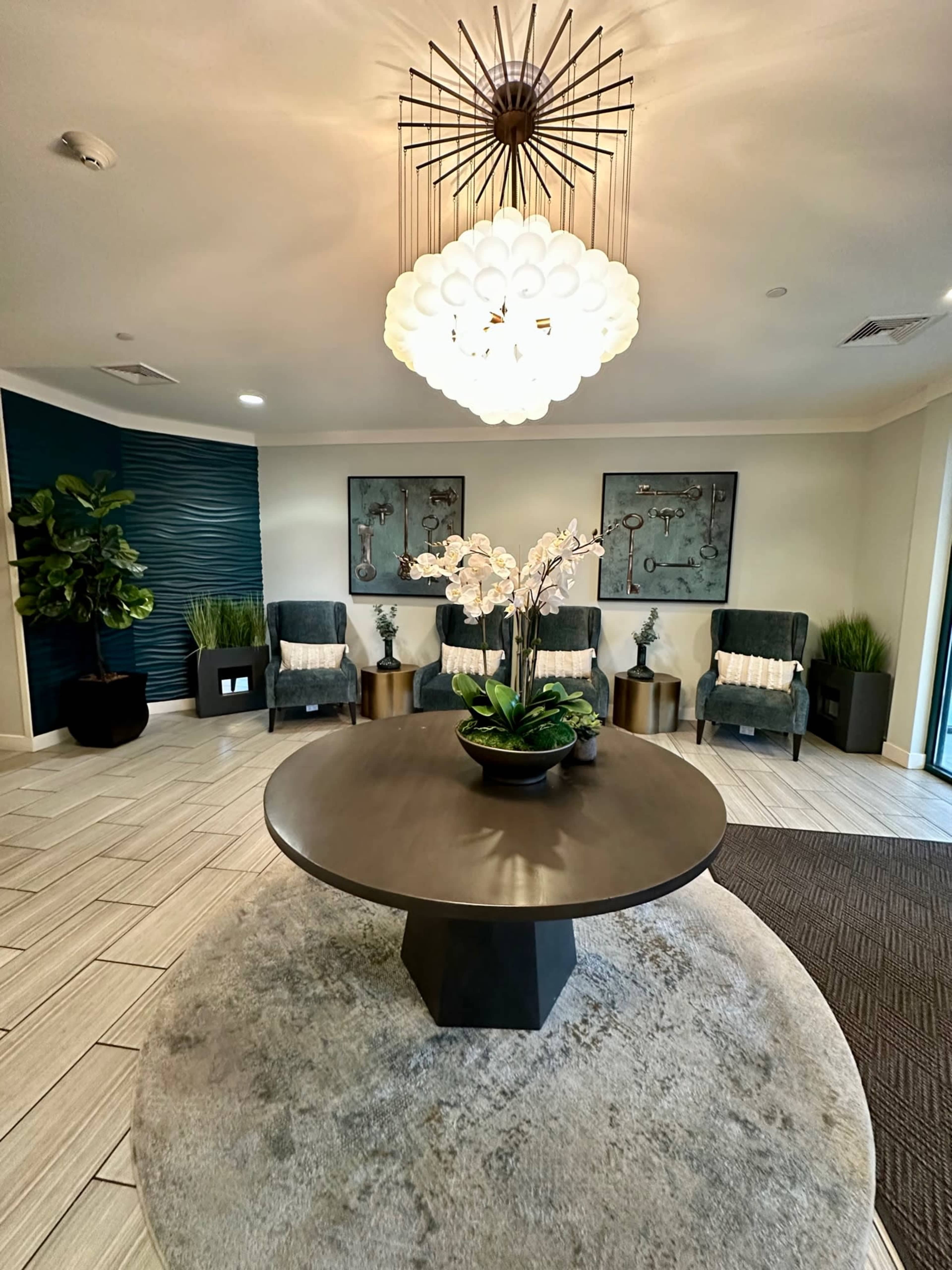 A stylish lobby area with a circular table at the center, surrounded by armchairs and decorative plants under a large light fixture.