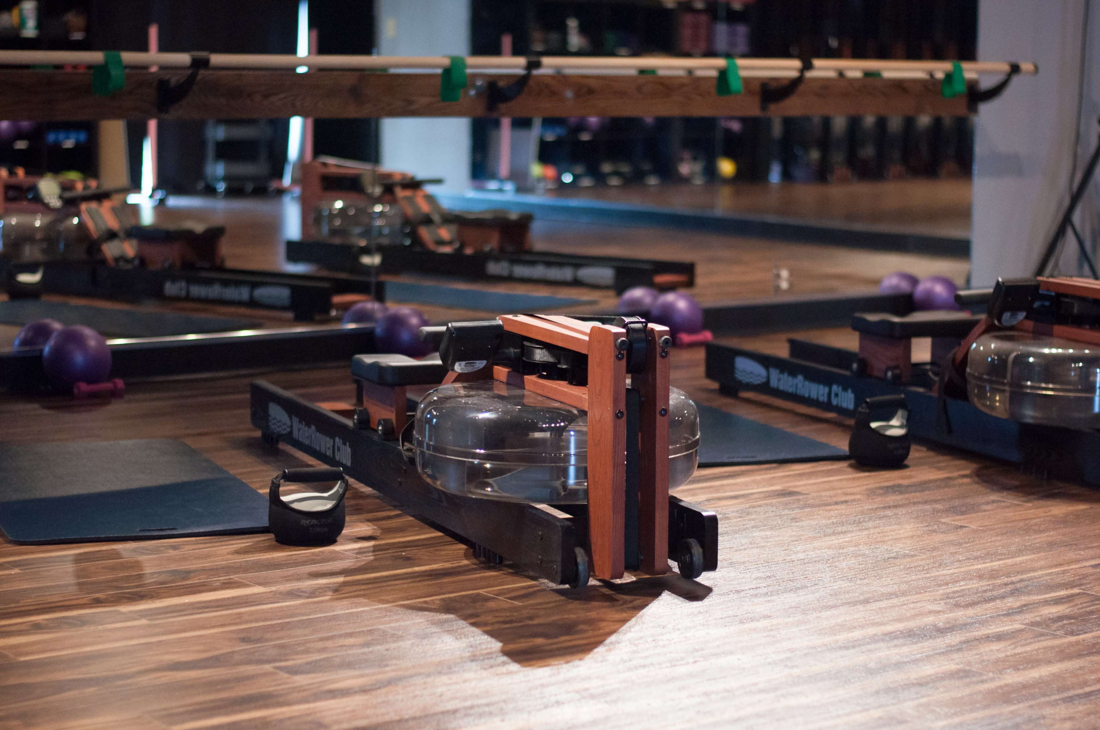 The image shows a wooden rowing machine with a water resistance tank in a fitness studio, surrounded by exercise mats and purple weights.