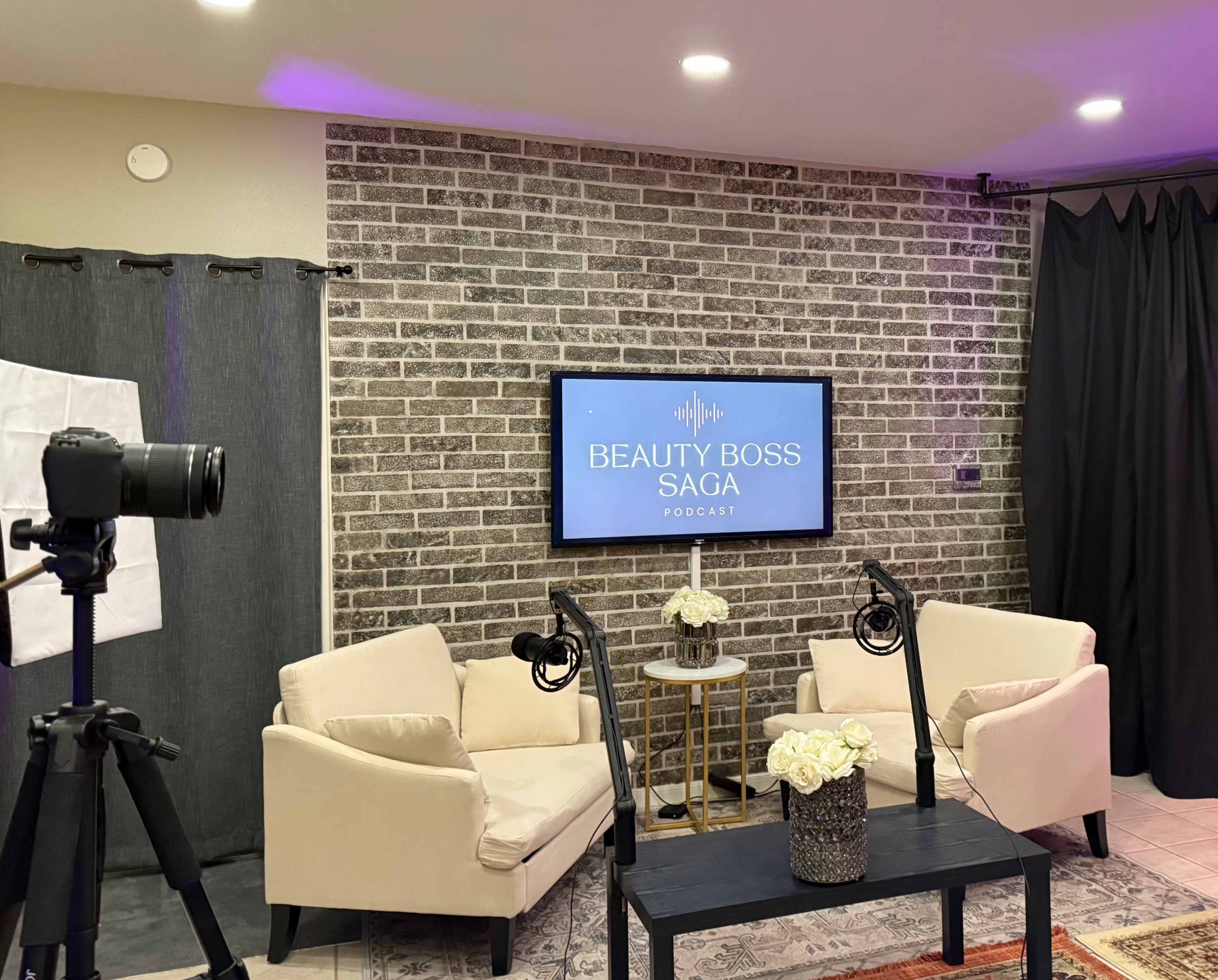 The image shows a podcast studio featuring two cream-colored chairs, a small table with flowers, a television displaying "BEAUTY BOSS SAGA PODCAST," and camera equipment set up nearby.