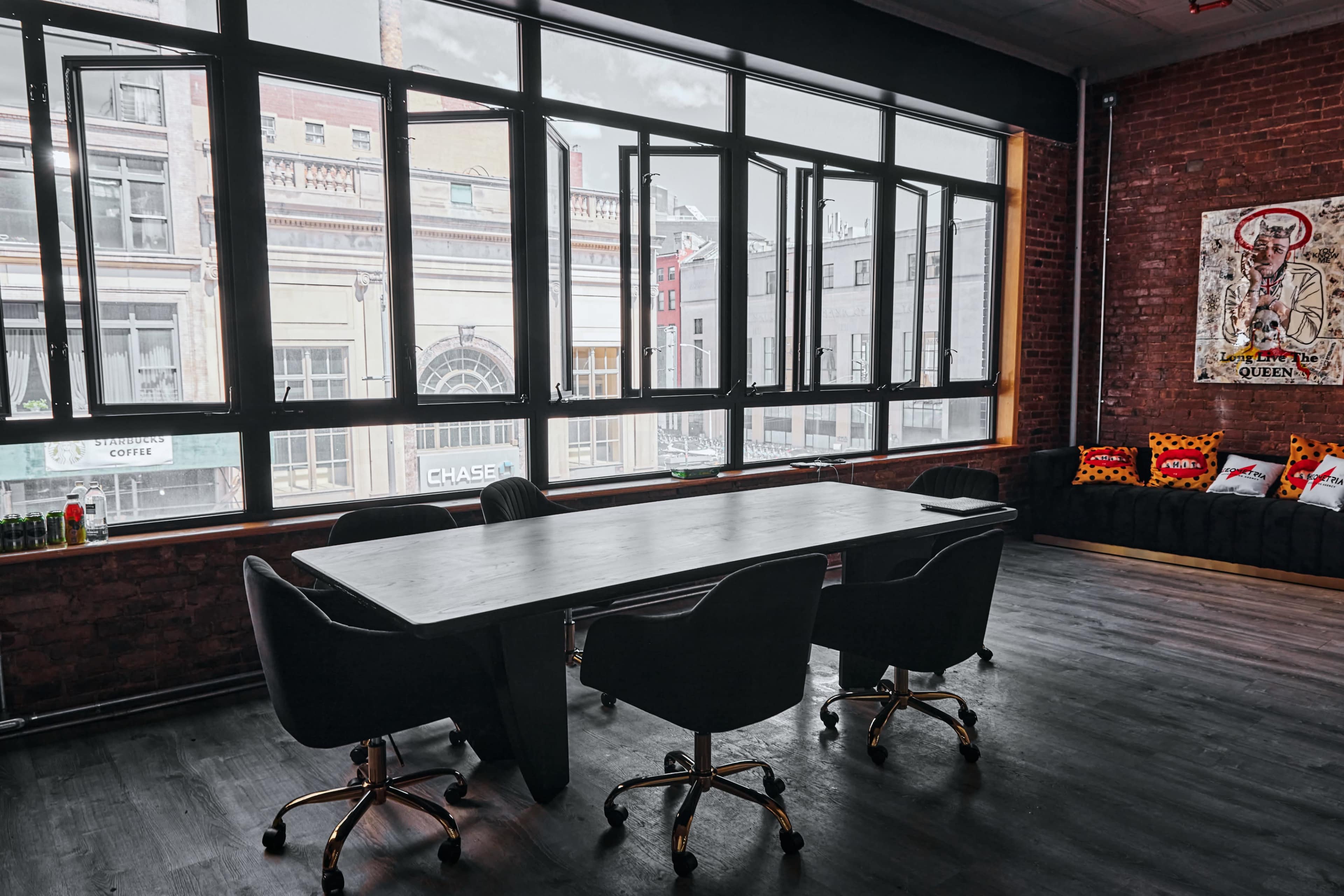 A modern conference room with a large table and four black chairs, featuring large windows that overlook a city street.