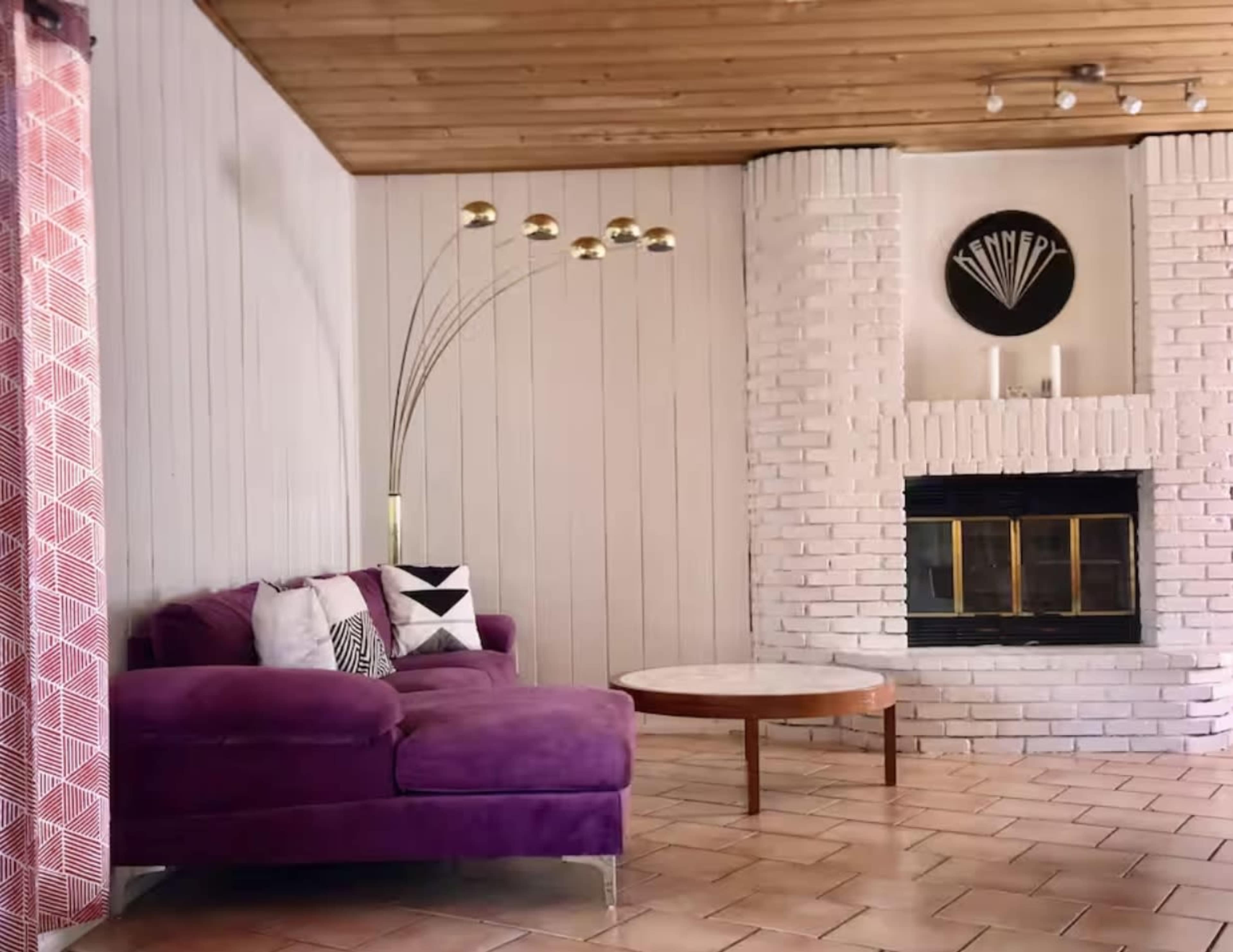 A purple sectional sofa is positioned next to a circular wooden coffee table in a room with a brick fireplace and wooden ceiling.