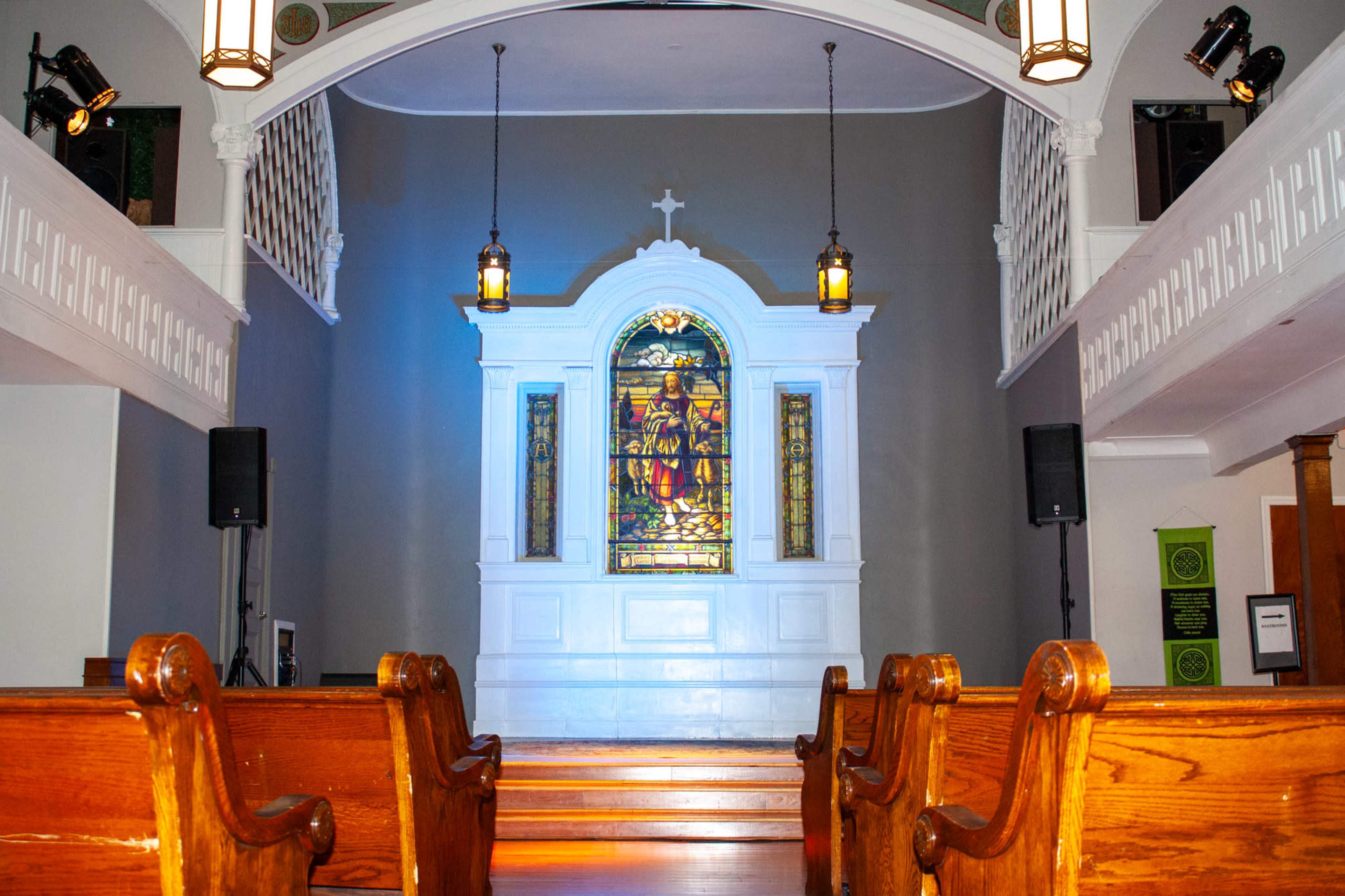 The interior of a church features wooden pews facing a stained glass window above an altar, illuminated by blue light.