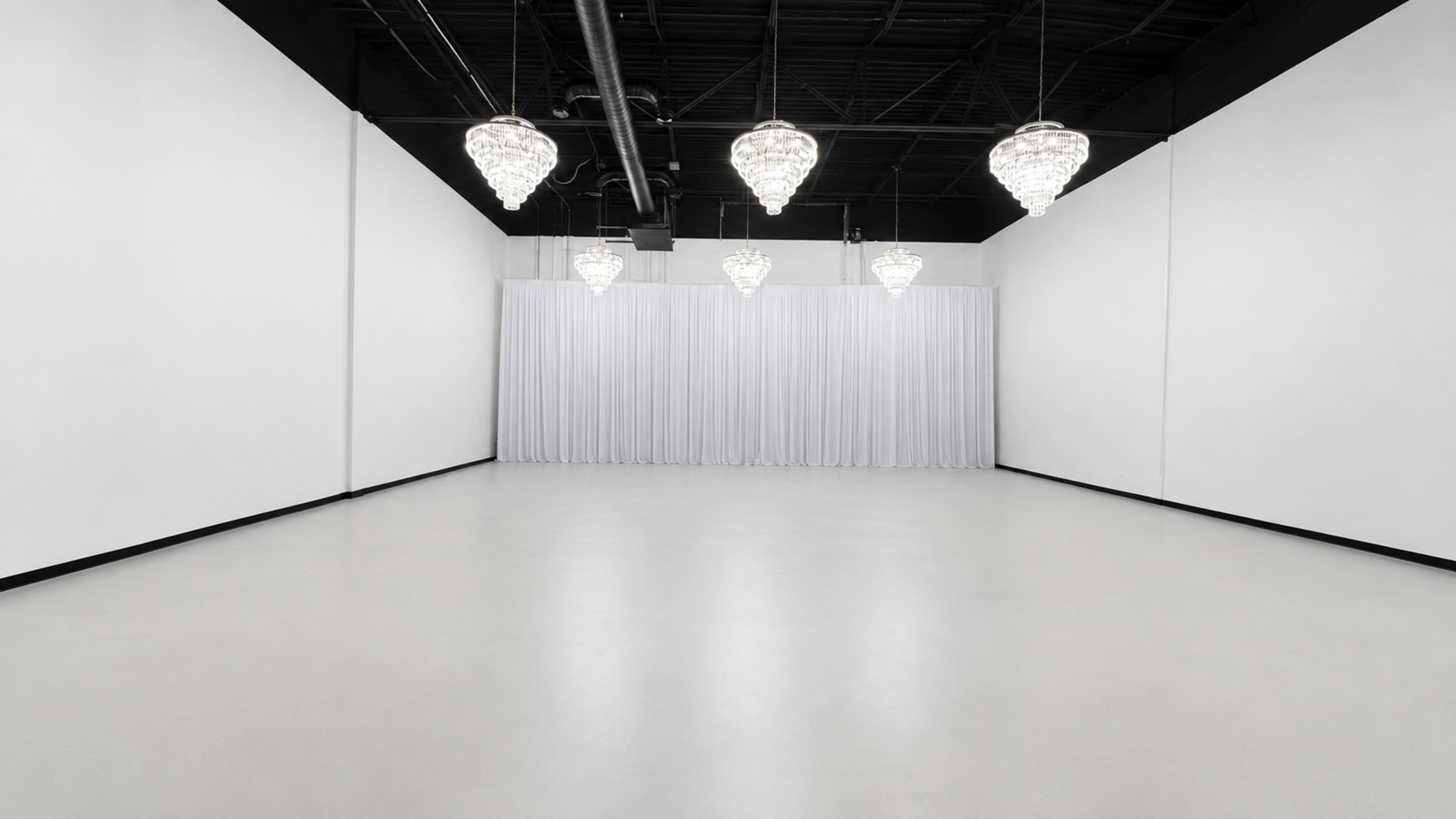 The image depicts a minimalist interior space with white walls, a light-colored floor, and several hanging chandeliers illuminating the area.