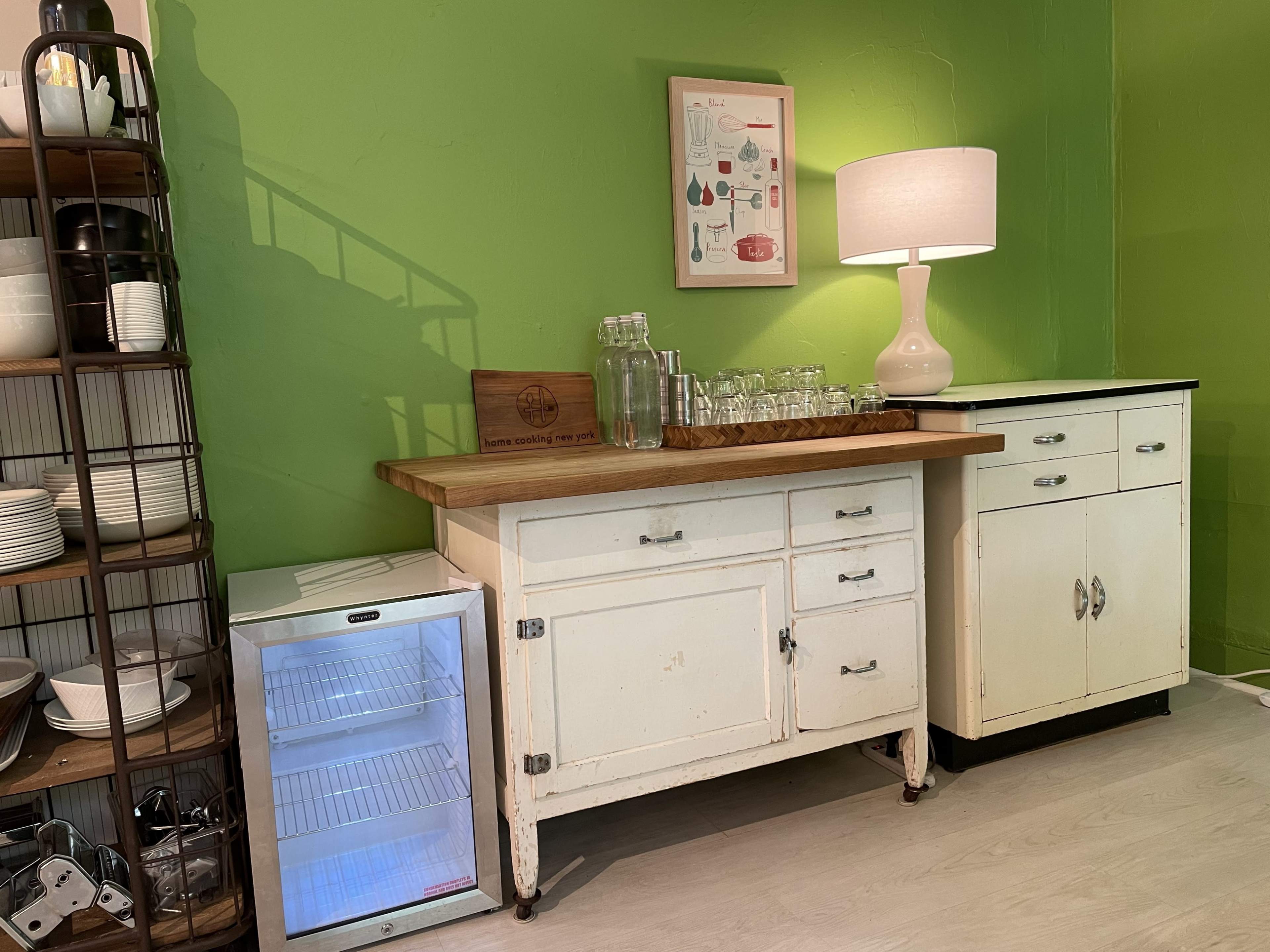 A kitchen area with a green wall, a wooden countertop with glassware, a small refrigerator, and a white sideboard with a lamp on it.