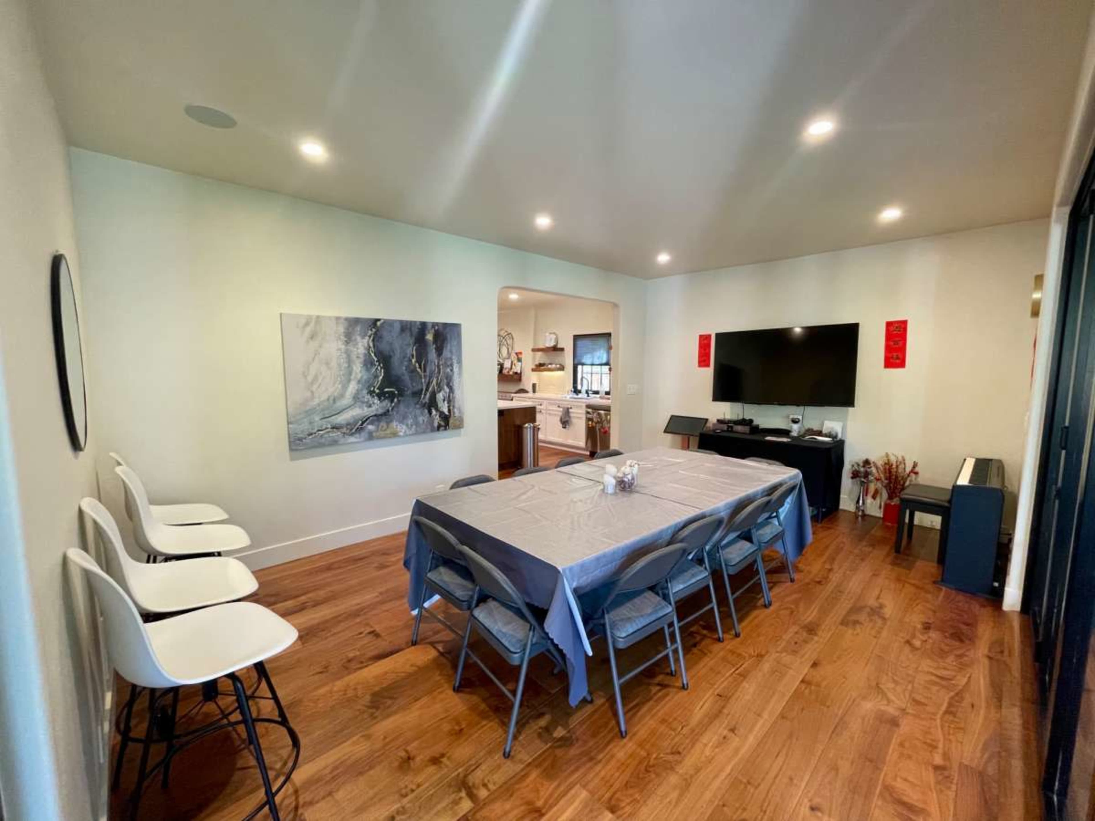 The image shows a modern dining room with a long rectangular table covered in a gray tablecloth, surrounded by folding chairs, and a TV mounted on the wall opposite a kitchen entrance.