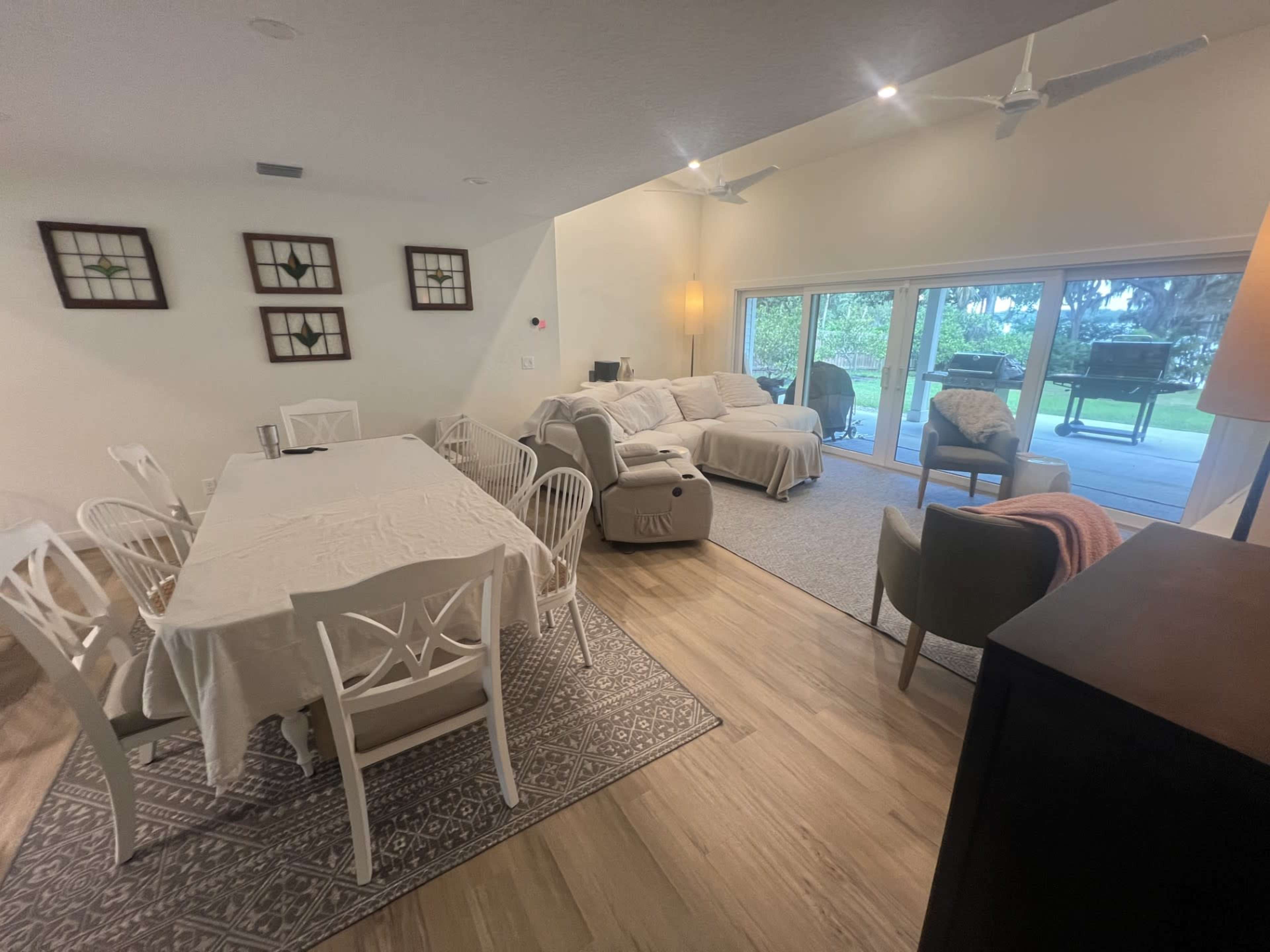 A spacious living area features a dining table with white chairs, a comfortable seating area, and large glass doors leading to an outdoor space.