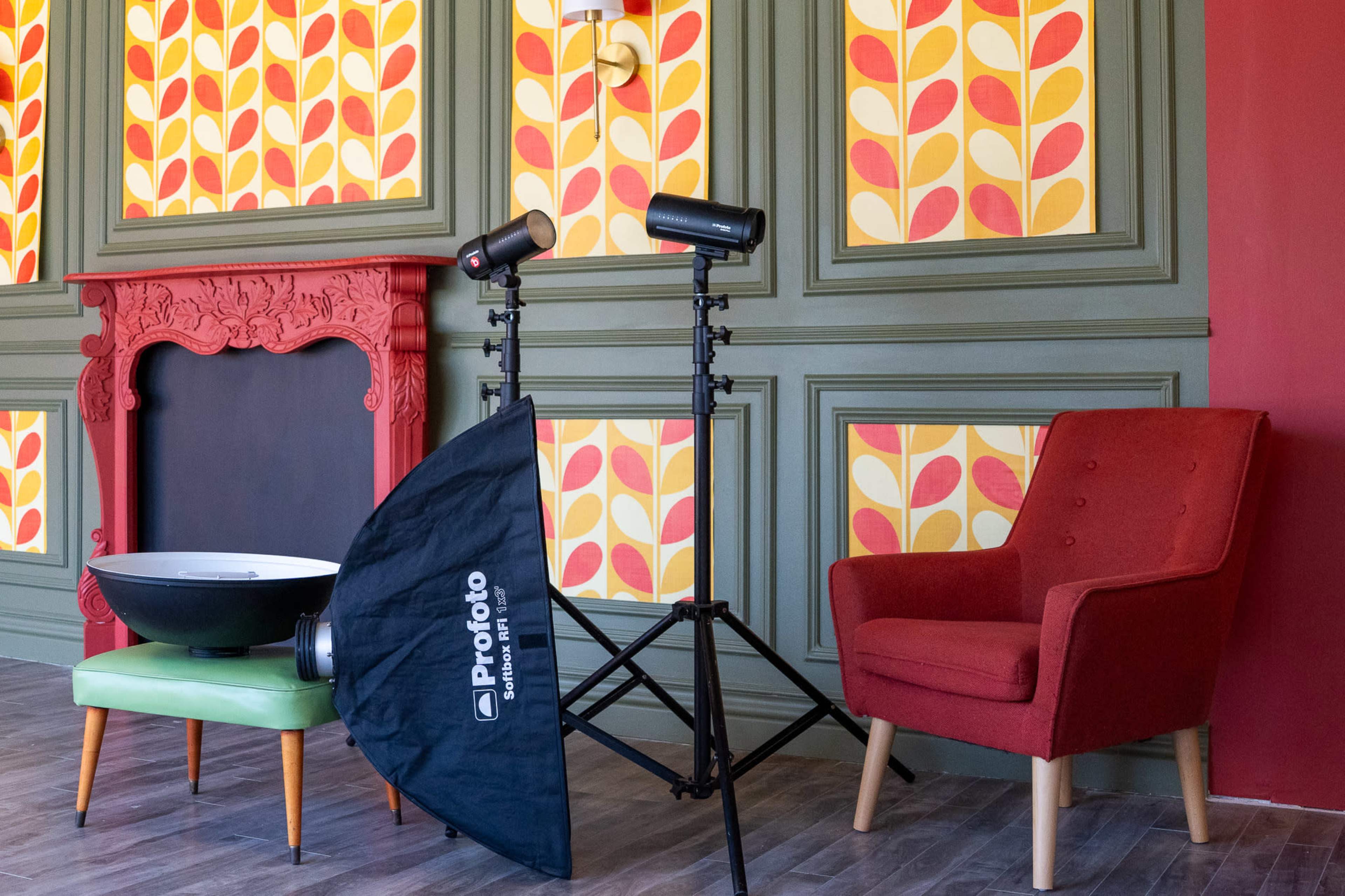 The scene features a photography setup with two lights on stands, a black softbox, a vintage-style red chair, and a decorative wall with colorful patterned wallpaper.