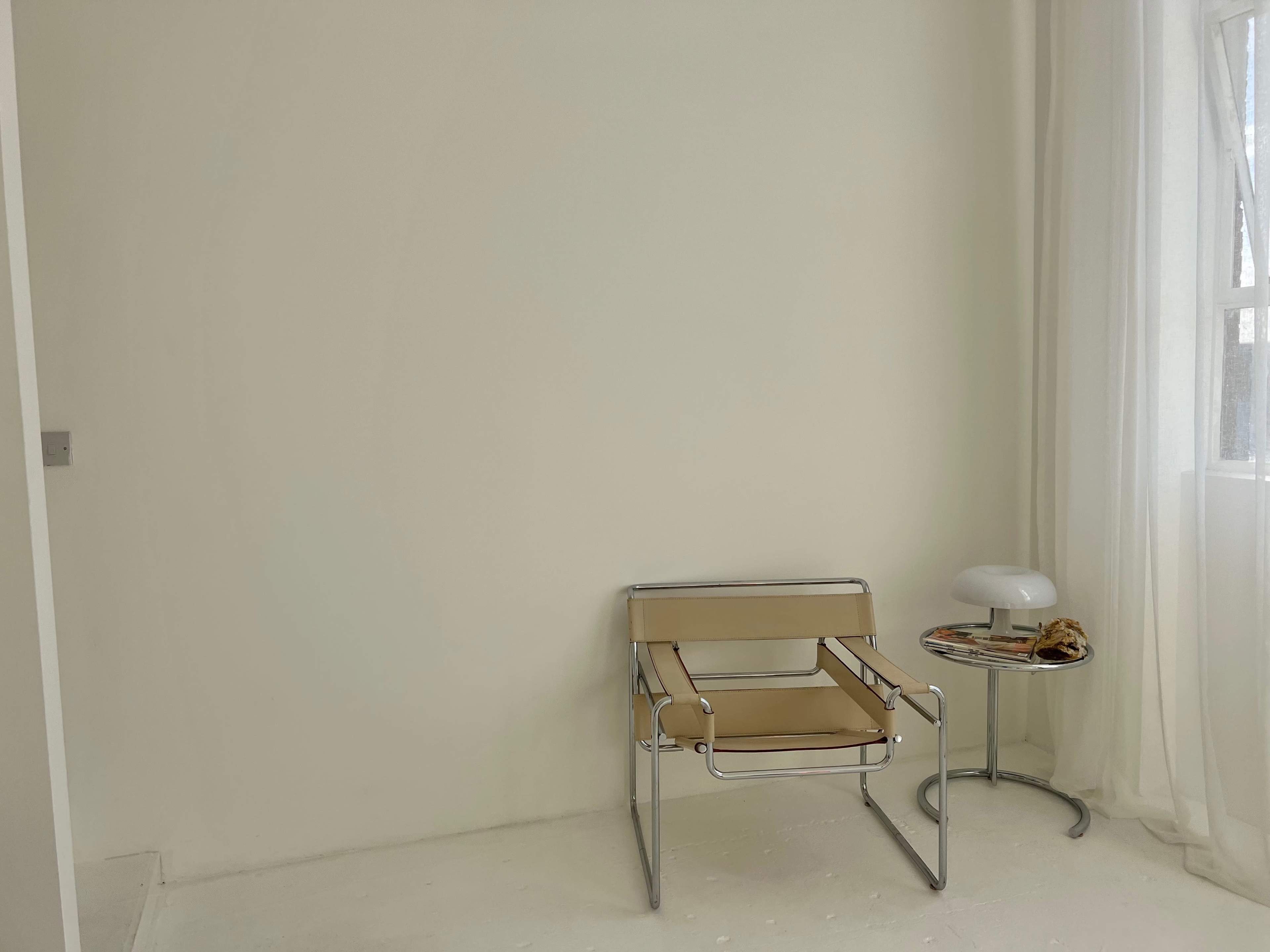 A minimalist interior features a light-colored wall, a chrome-framed chair with a tan seat, and a small side table with a lamp and magazines.