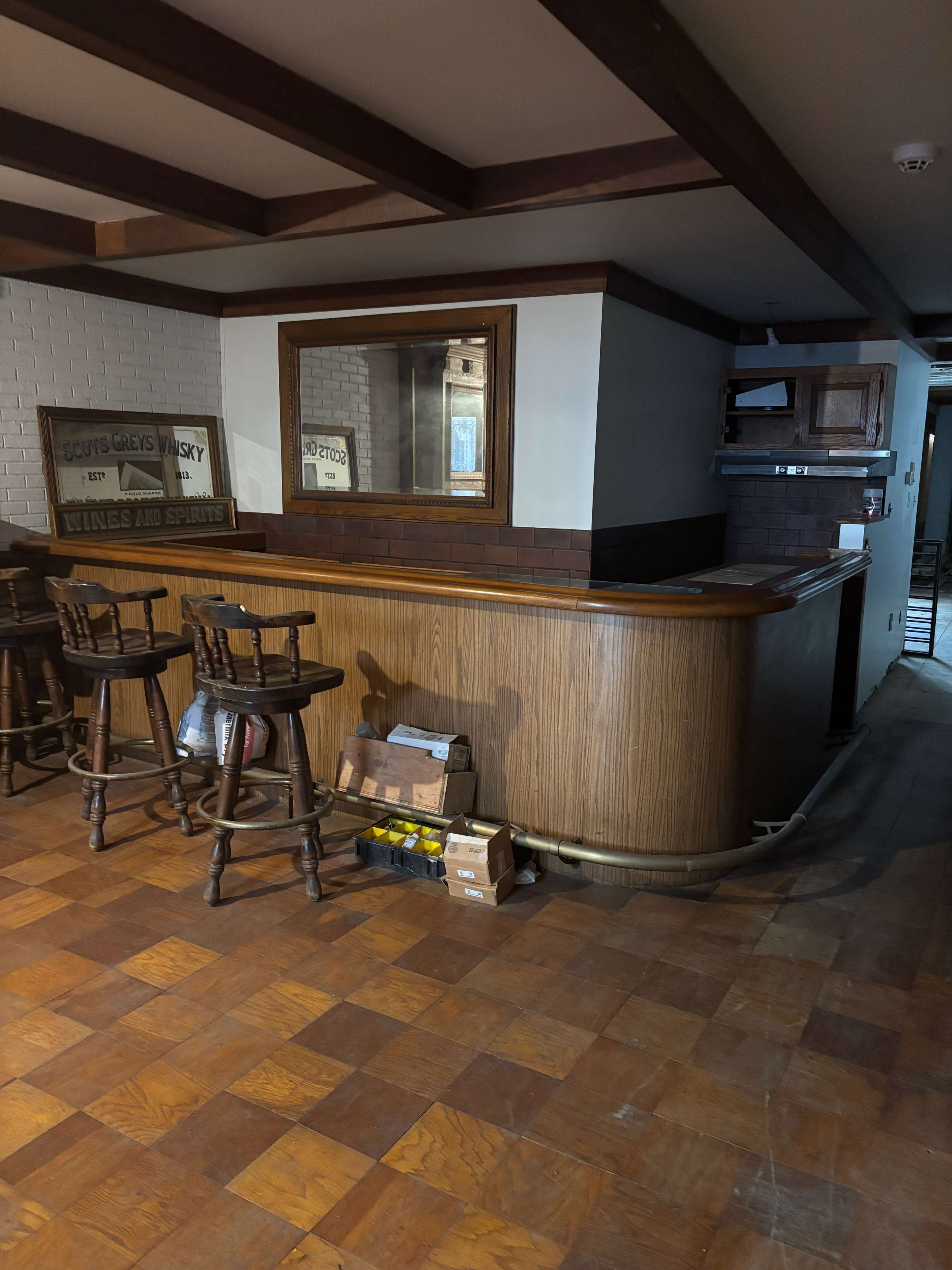 The image shows a wooden bar setup with high stools, situated in a spacious room with tiled flooring and exposed wooden beams.