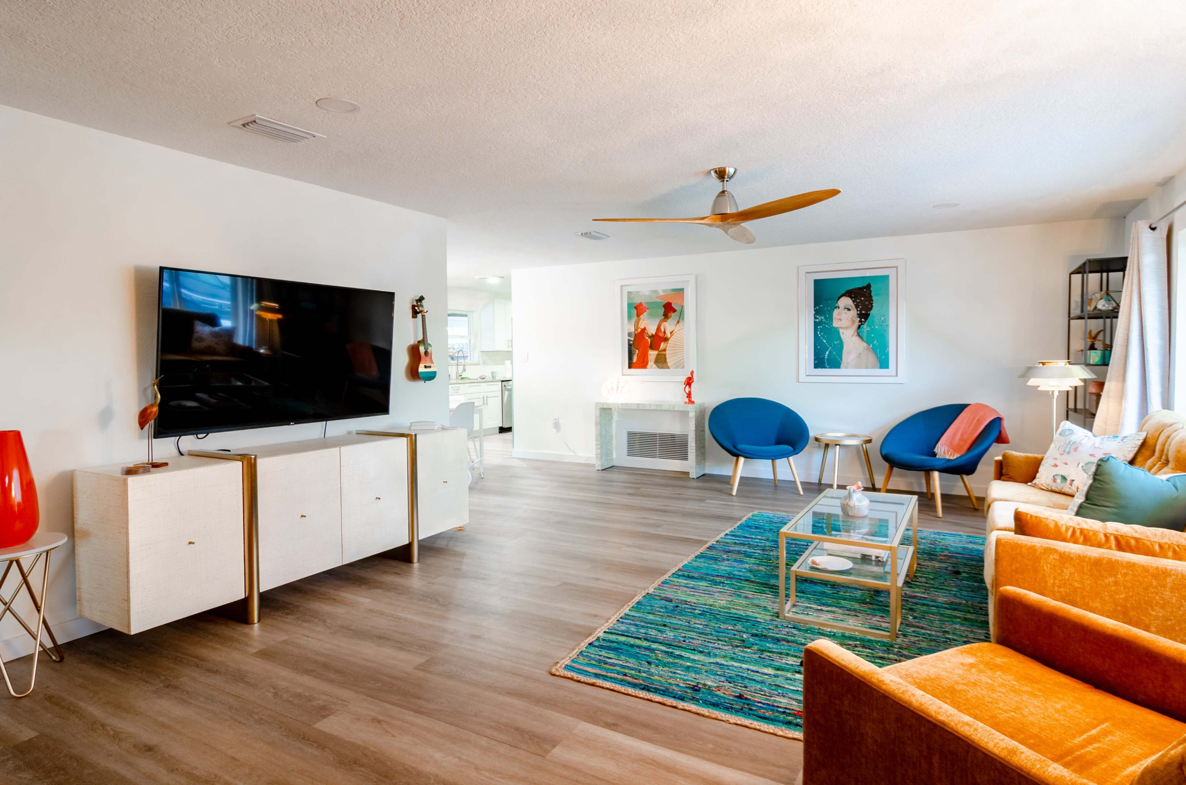 A modern living room featuring a large television mounted on the wall, a glass coffee table, colorful chairs, and a bright area rug.