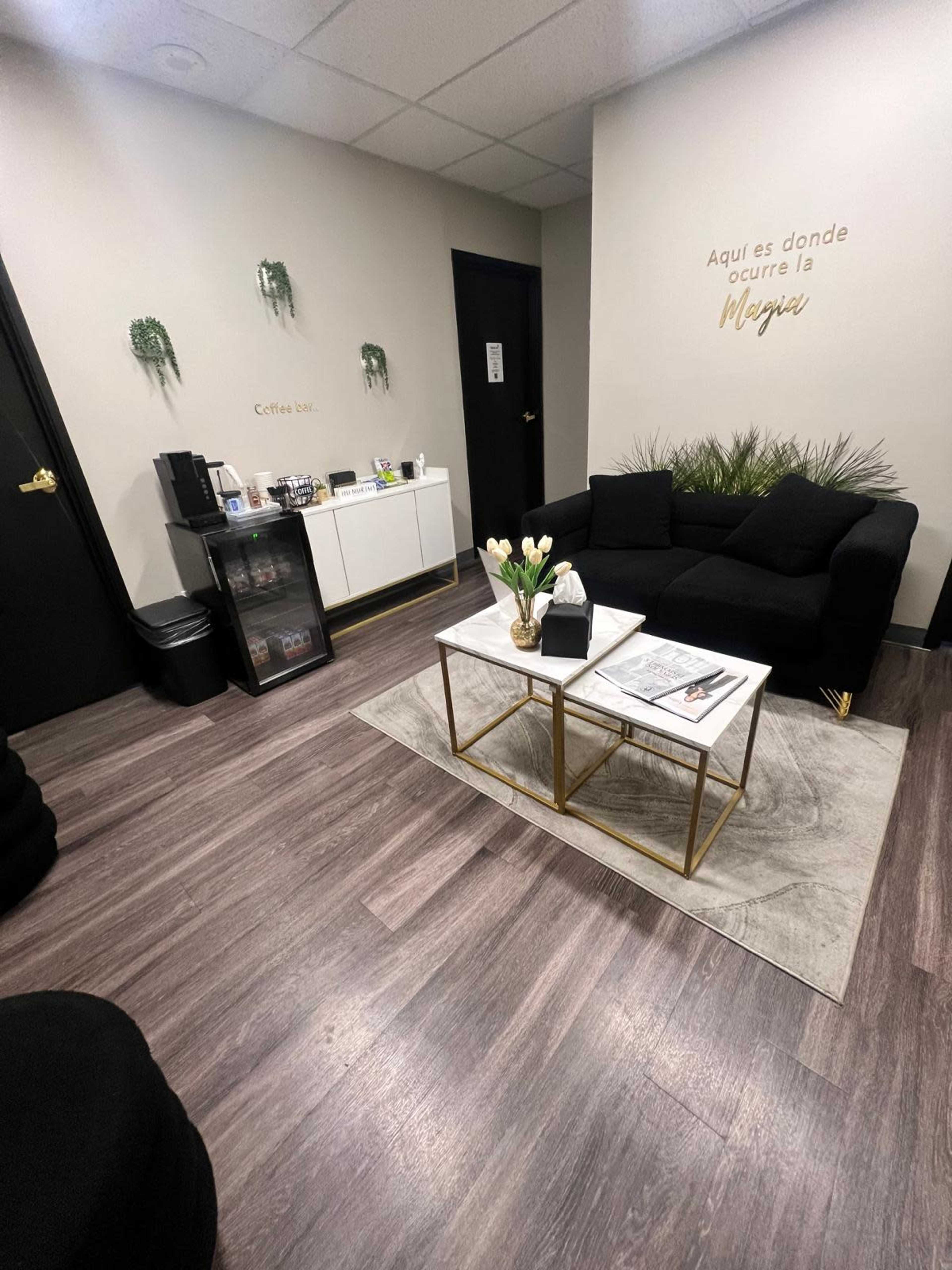 The image shows a modern waiting room with black seating, a coffee station, and decorative plants, featuring stylish furniture and neutral wall colors.