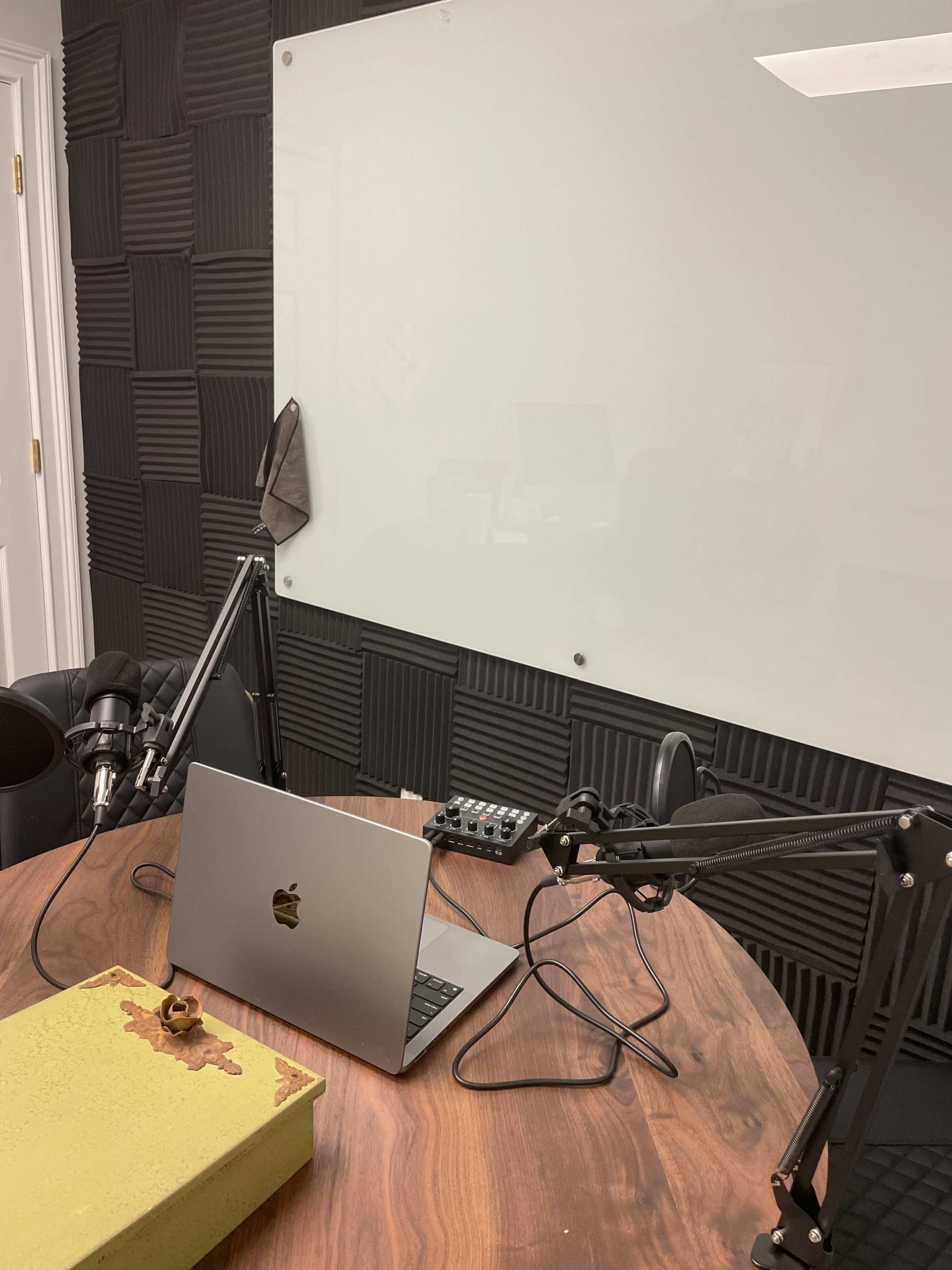 The image shows a recording studio setup with a wooden table, a laptop, microphones mounted on adjustable arms, and a soundproofed wall featuring black acoustic panels.