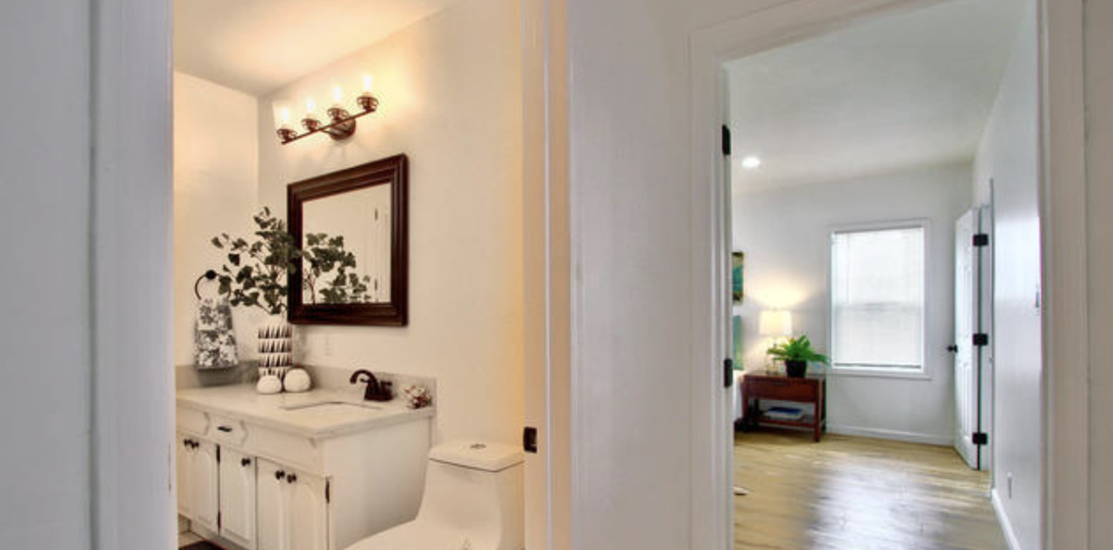 The image shows a bathroom on the left with a vanity, mirror, and toilet, and a hallway leading to a living area on the right, featuring a window and a piece of furniture.