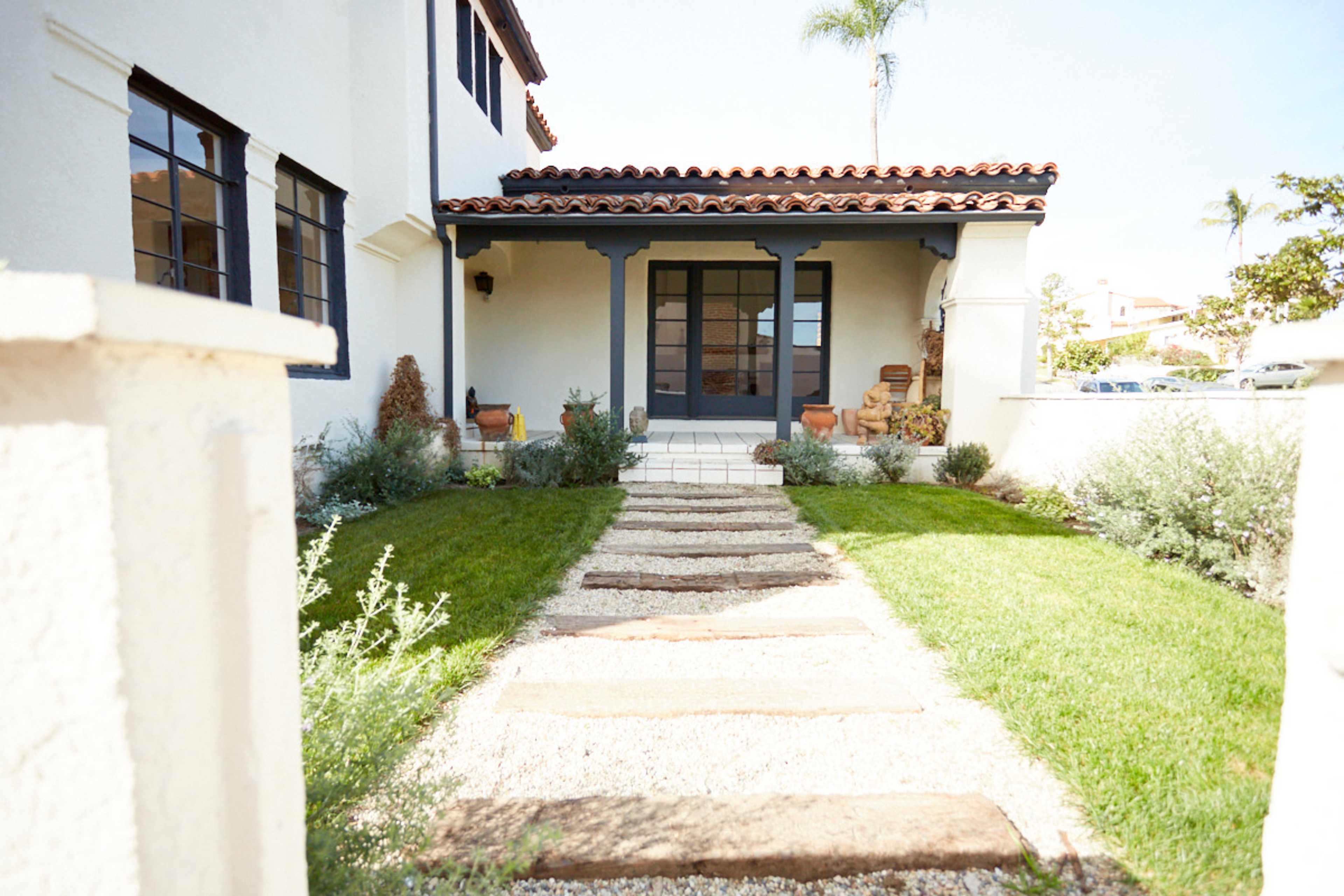 The image shows a well-maintained pathway leading to the entrance of a house, bordered by grass and decorative plants.