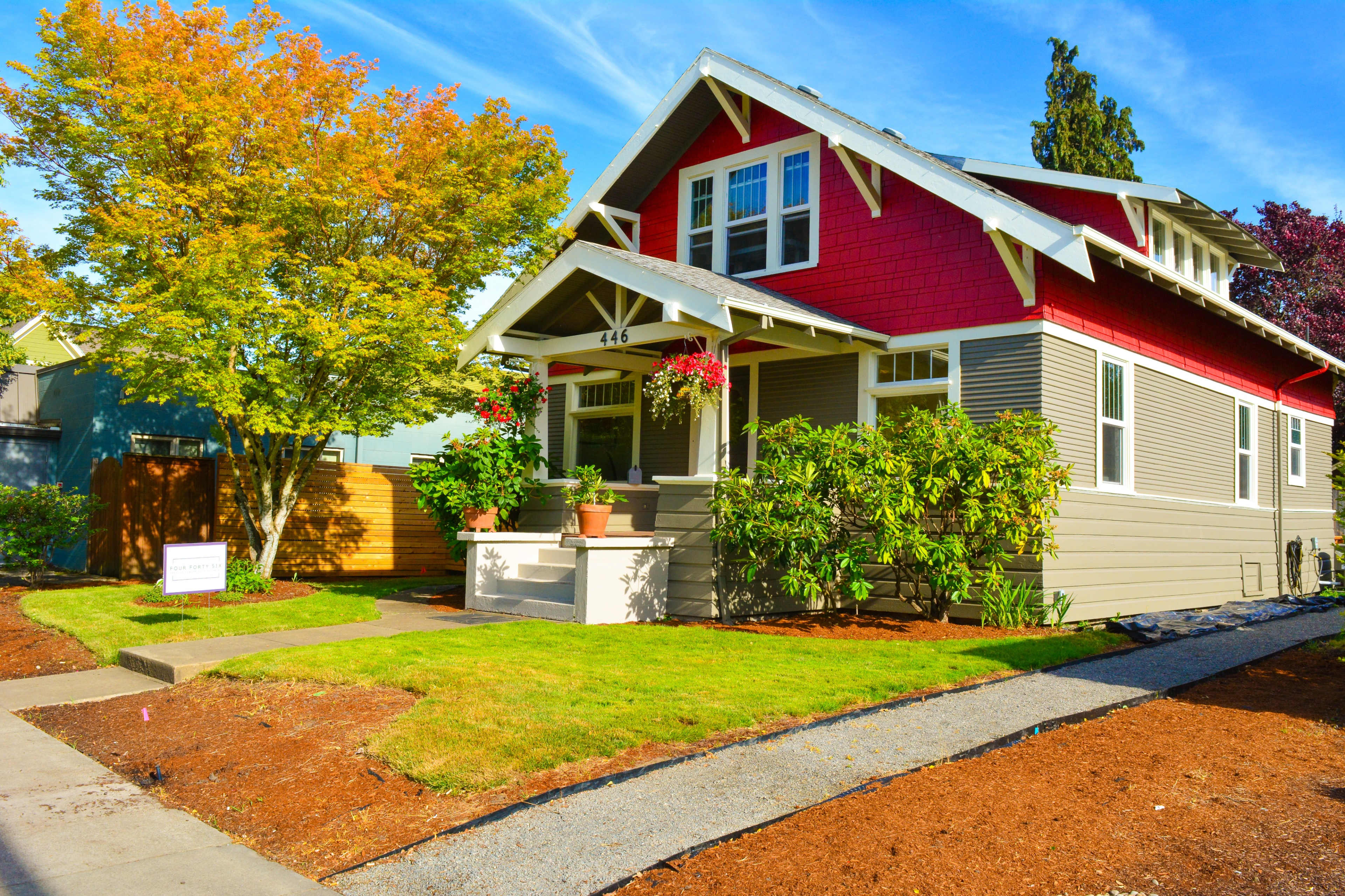 A two-story house with a red and gray exterior, surrounded by green plants and a well-maintained lawn, stands on a corner lot.