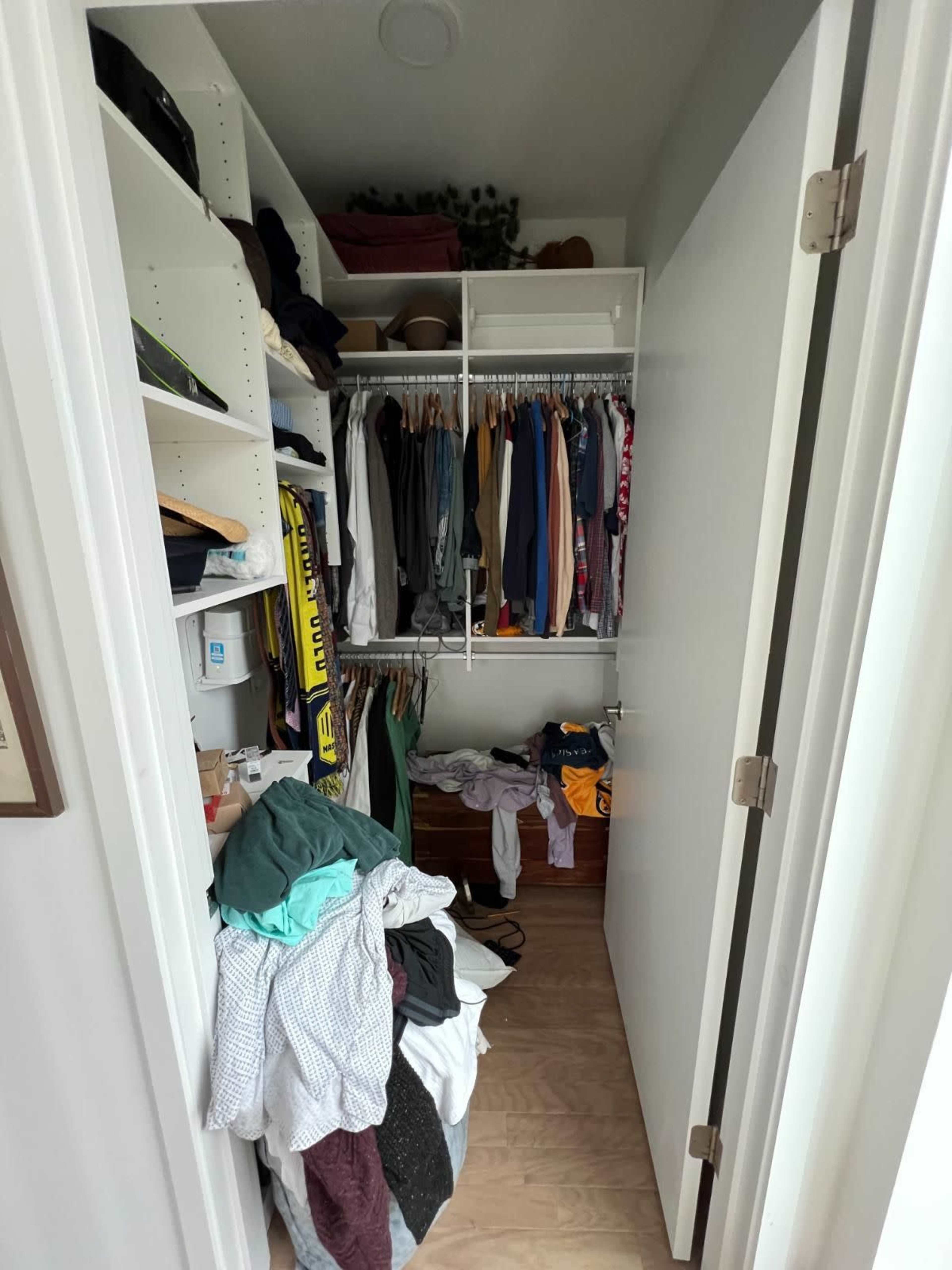 A cluttered closet with clothing piled on the floor and hanging on shelves and a rod.