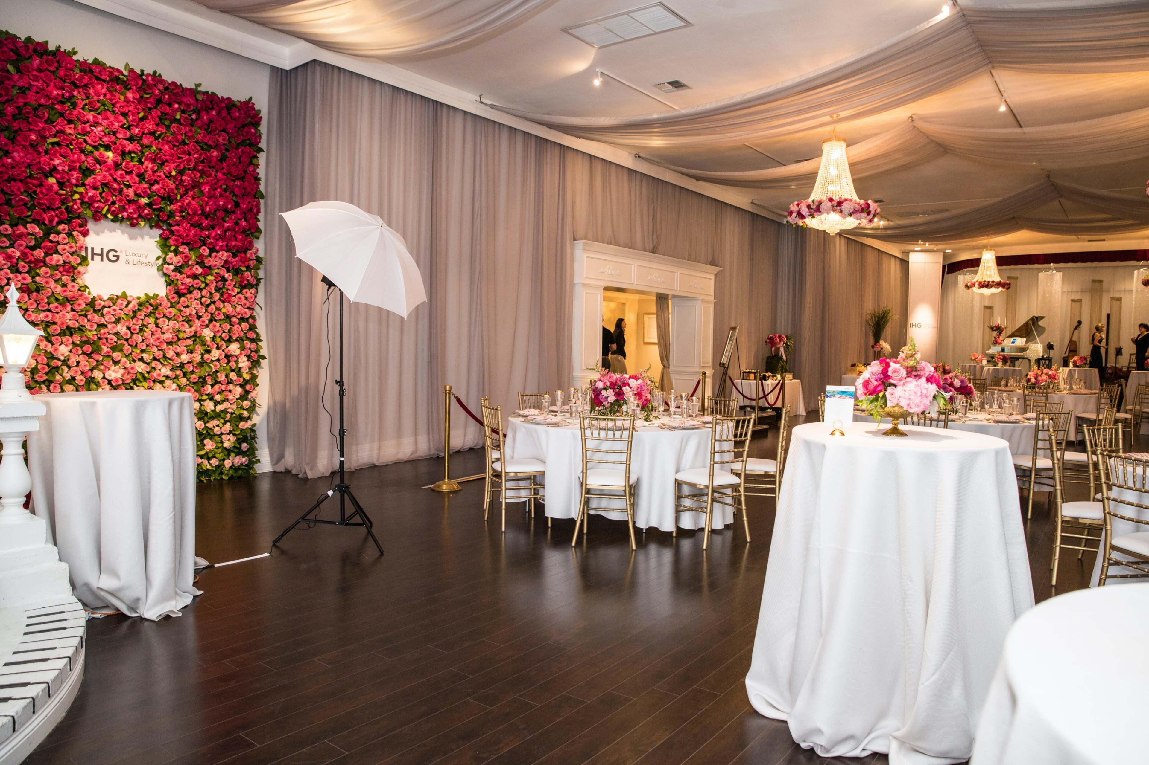 A spacious event venue features elegantly set tables, a vibrant floral backdrop, and soft draperies overhead.