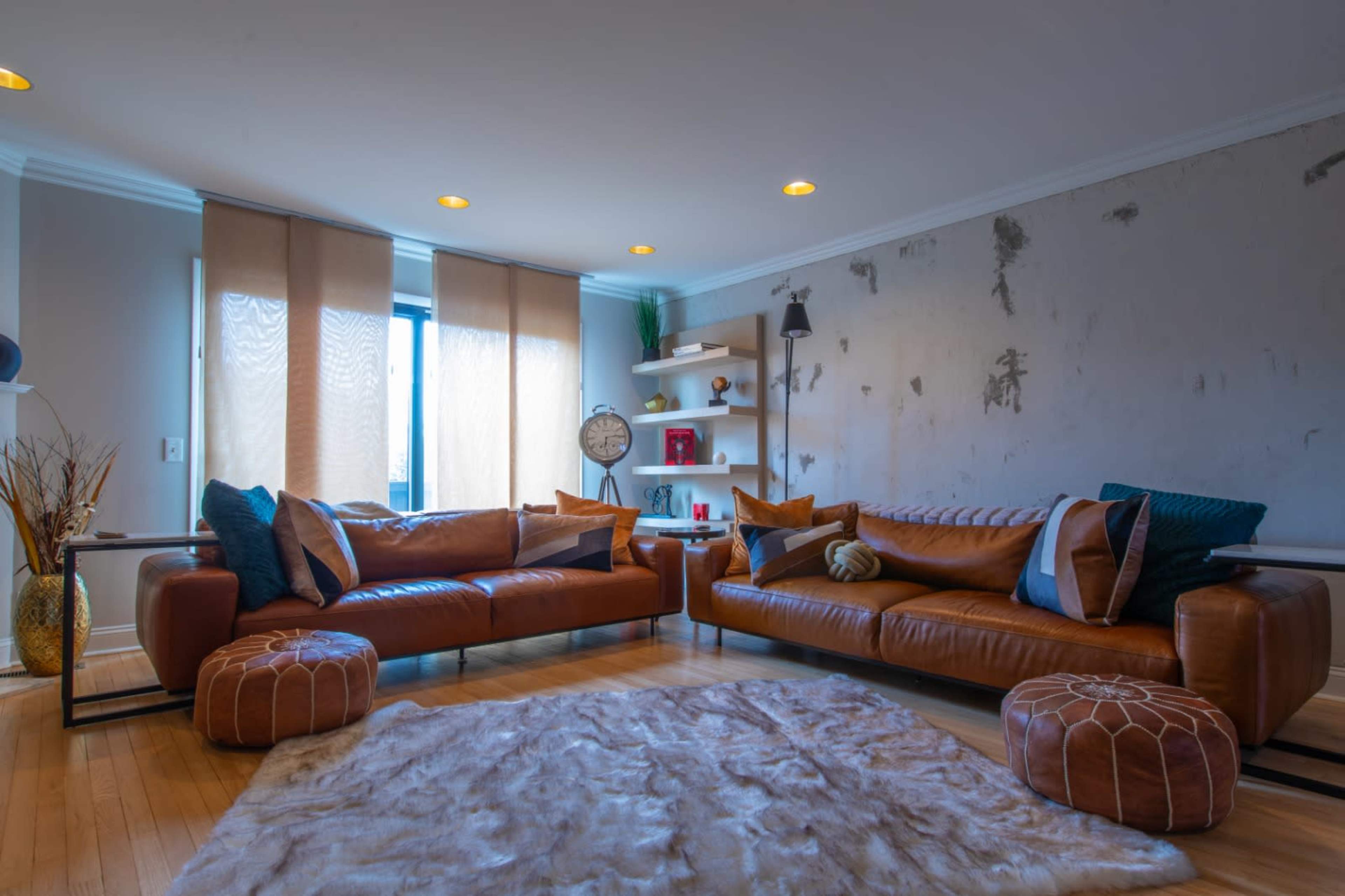 A modern living room features two leather sofas arranged opposite each other, with decorative pillows, a plush rug, wall shelves, and natural light from large windows.