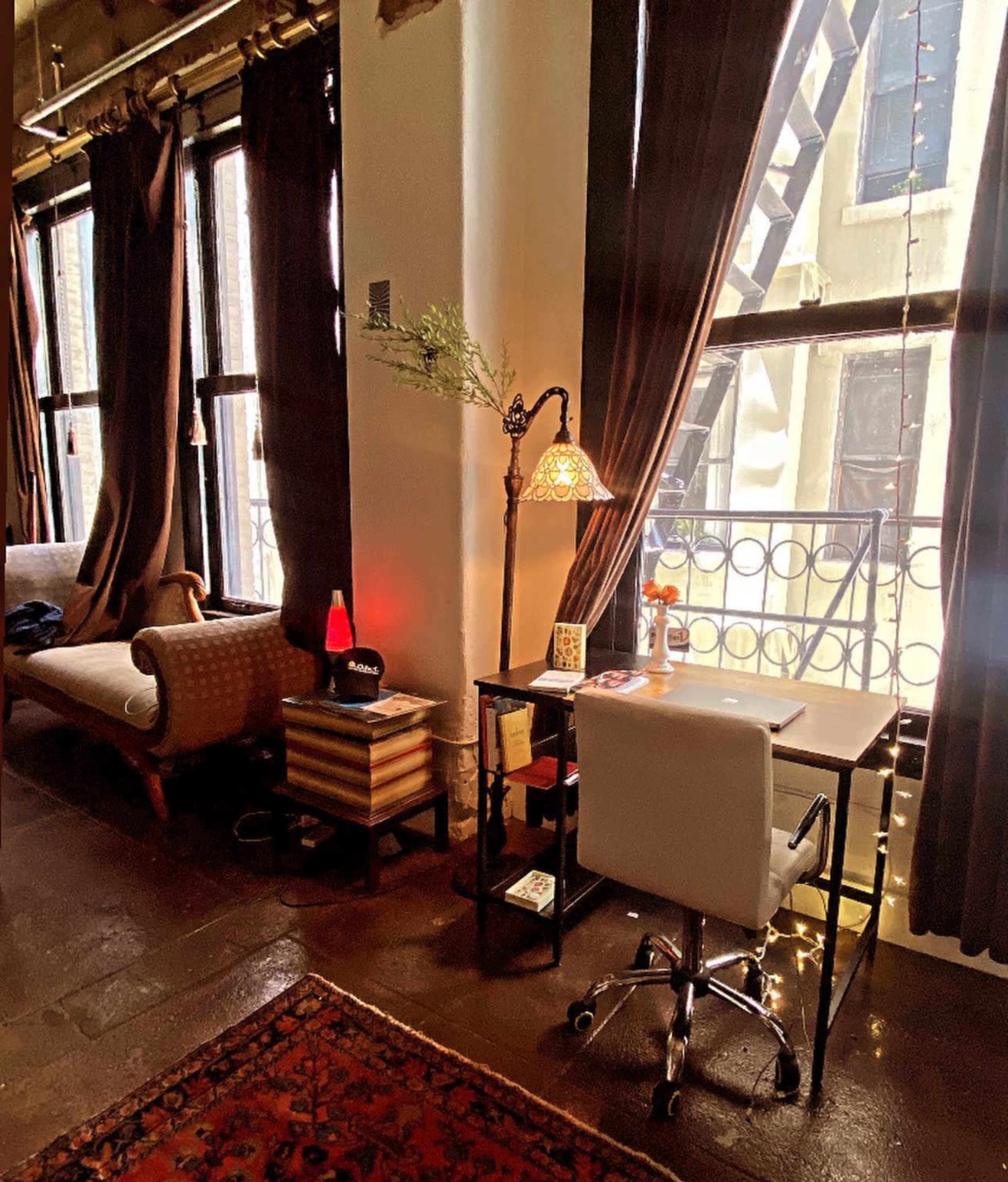 The image shows a cozy workspace with a small desk and chair, a lamp, and a decorative tree branch, adjacent to a sofa and large windows dressed with dark curtains.